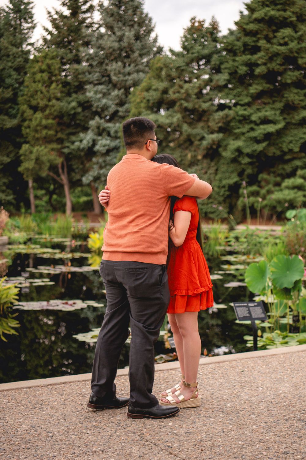 Two figures stand by a tranquil pond, one wearing orange and the other in a vibrant red dress, surrounded by evergreens.