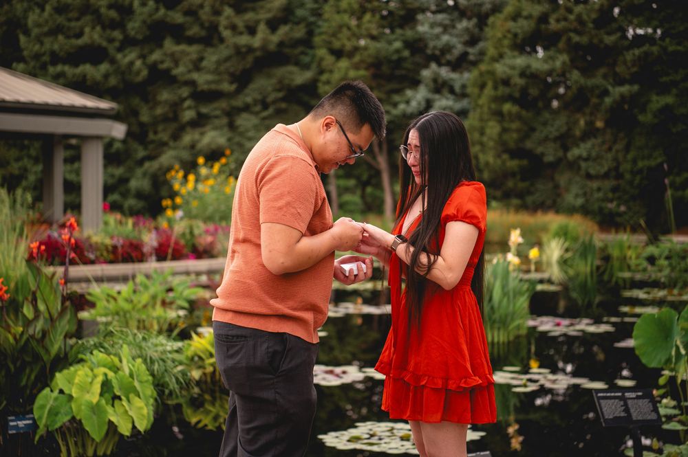 A couple in orange and red outfits stand together in a lush garden setting during a special occasion.