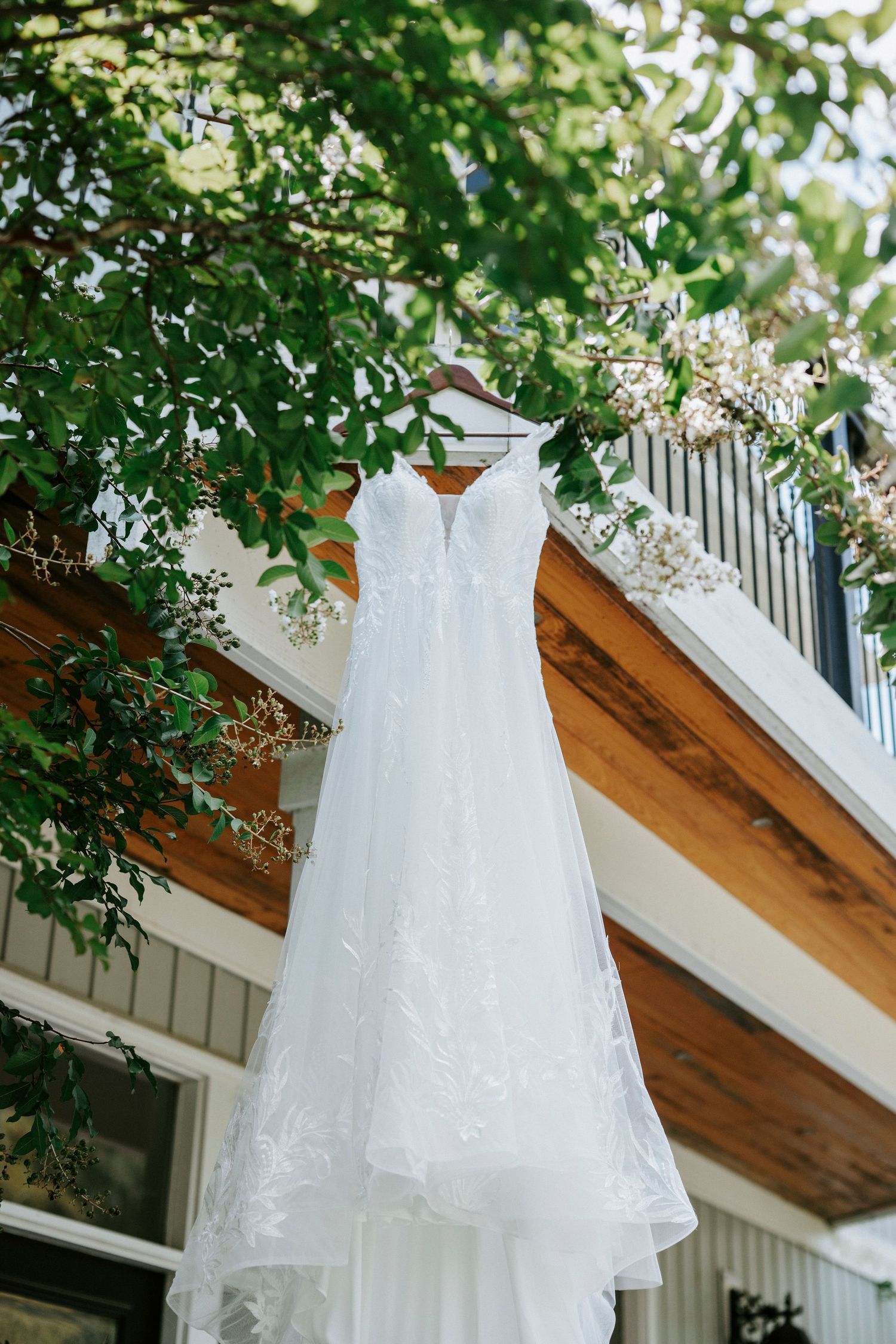 White wedding dress hanging outdoors against green foliage and wooden architecture.