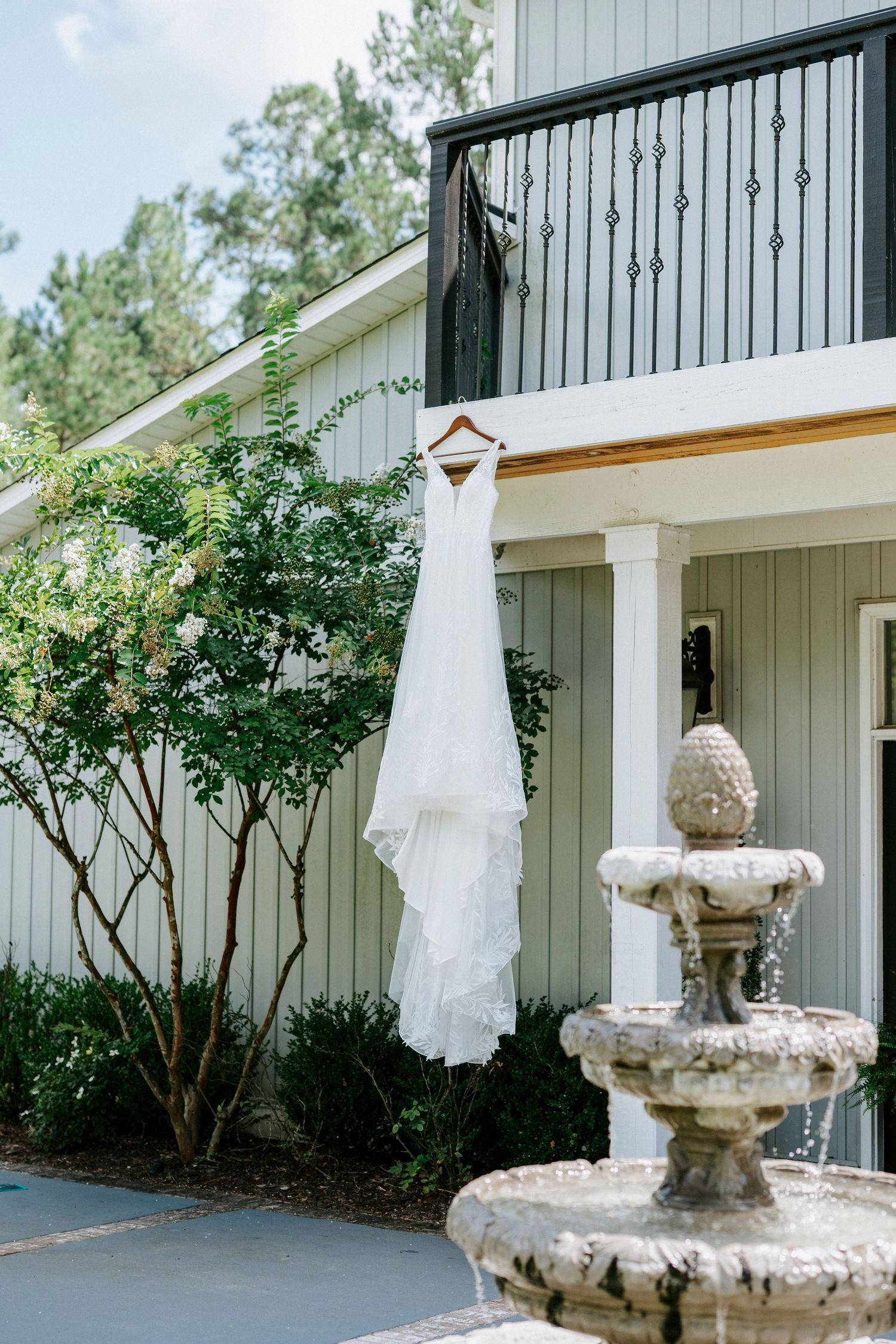 White wedding dress hanging on exterior of rustic white barn building with stone fountain in foreground.