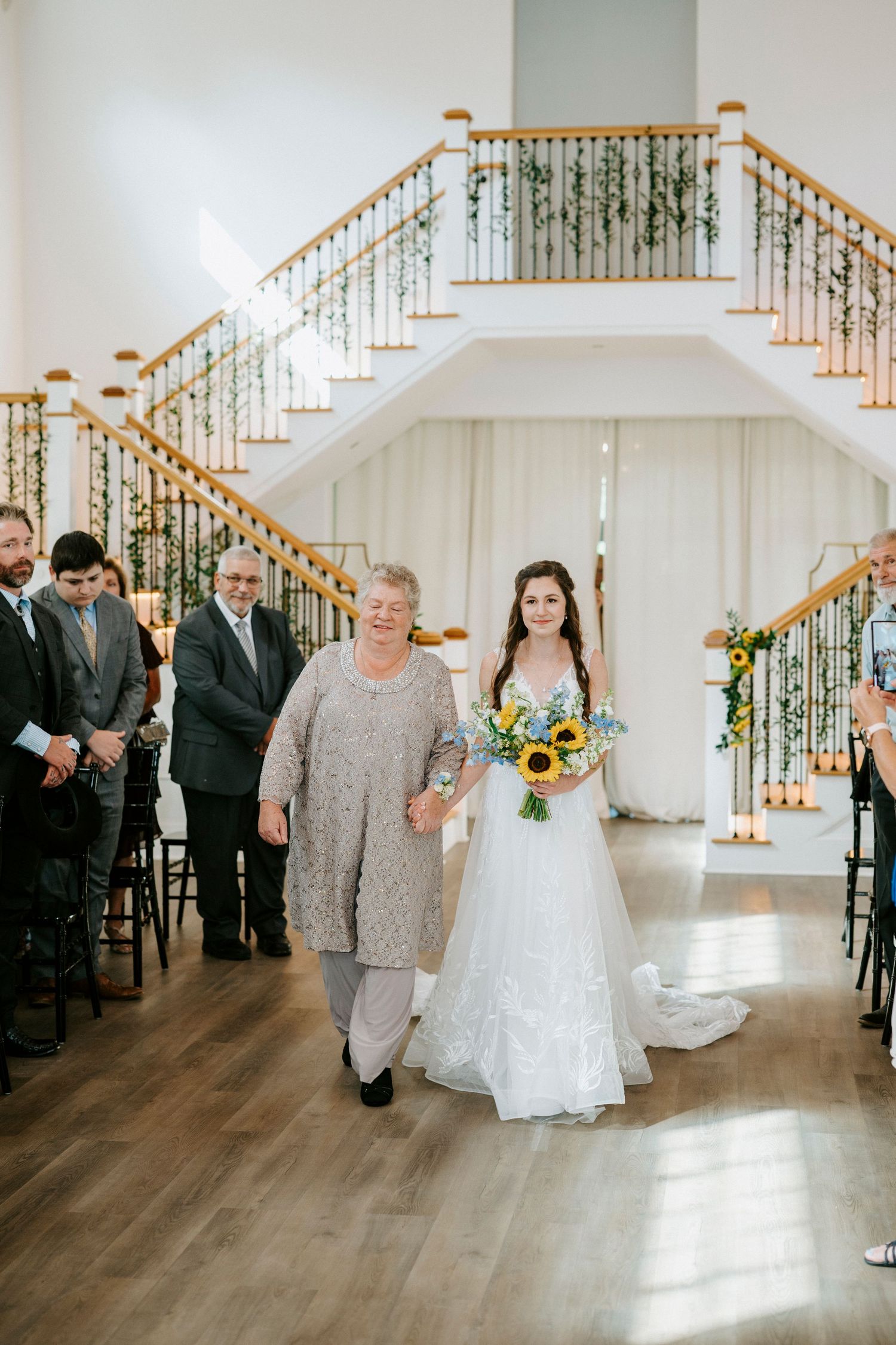 A bride in white carrying sunflowers walks down the aisle during a wedding ceremony.