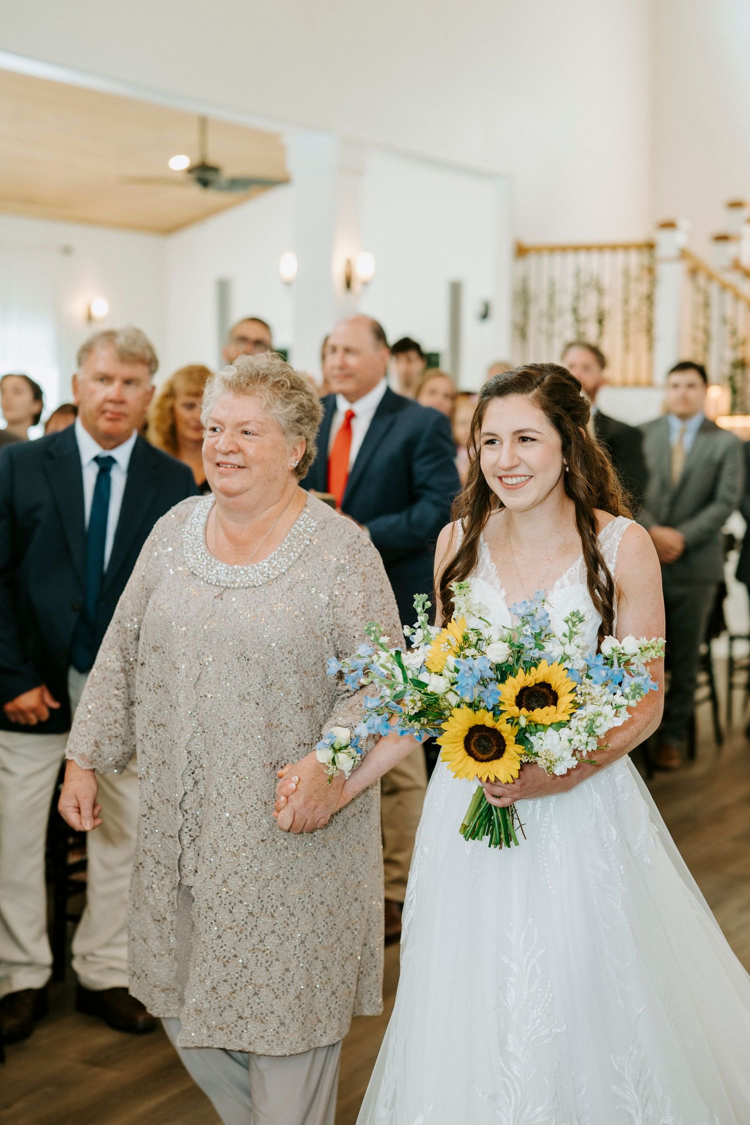 Wedding ceremony processional with attendants carrying sunflower bouquets in an elegant indoor venue.