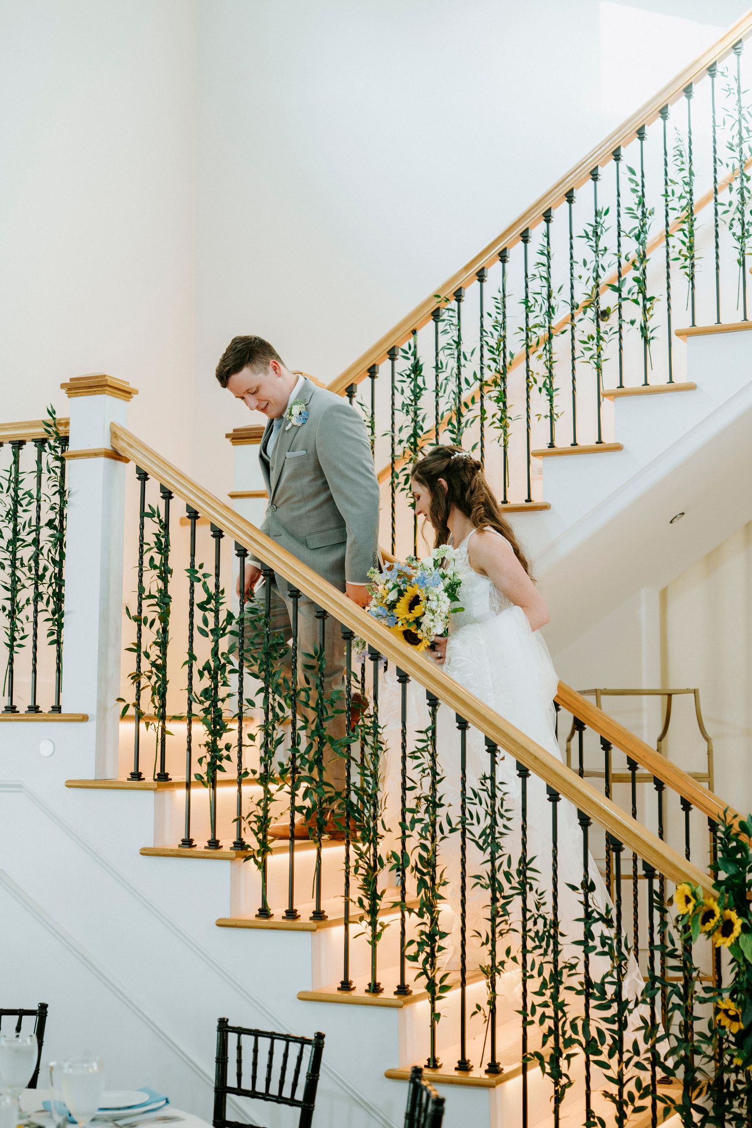 A bride and groom descend an elegant staircase decorated with greenery and wooden railings.