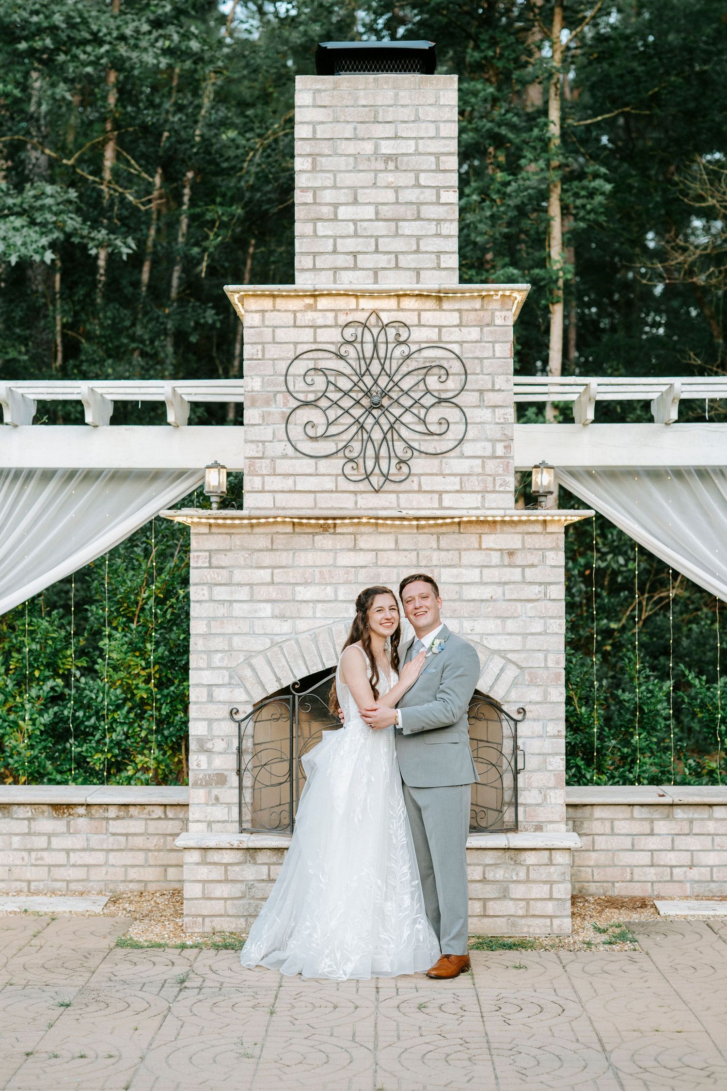 Wedding couple embraces in front of white brick pillars and draped fabric at outdoor garden venue.