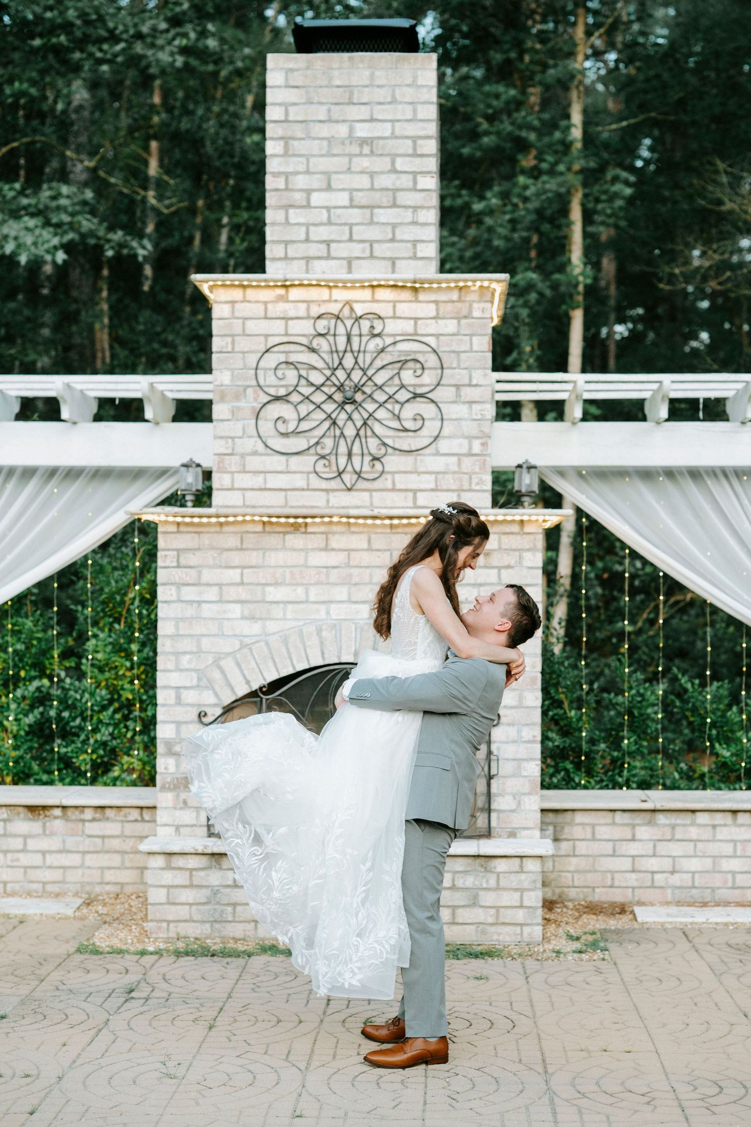 A groom lifts his bride in a romantic pose in front of an outdoor brick fireplace.