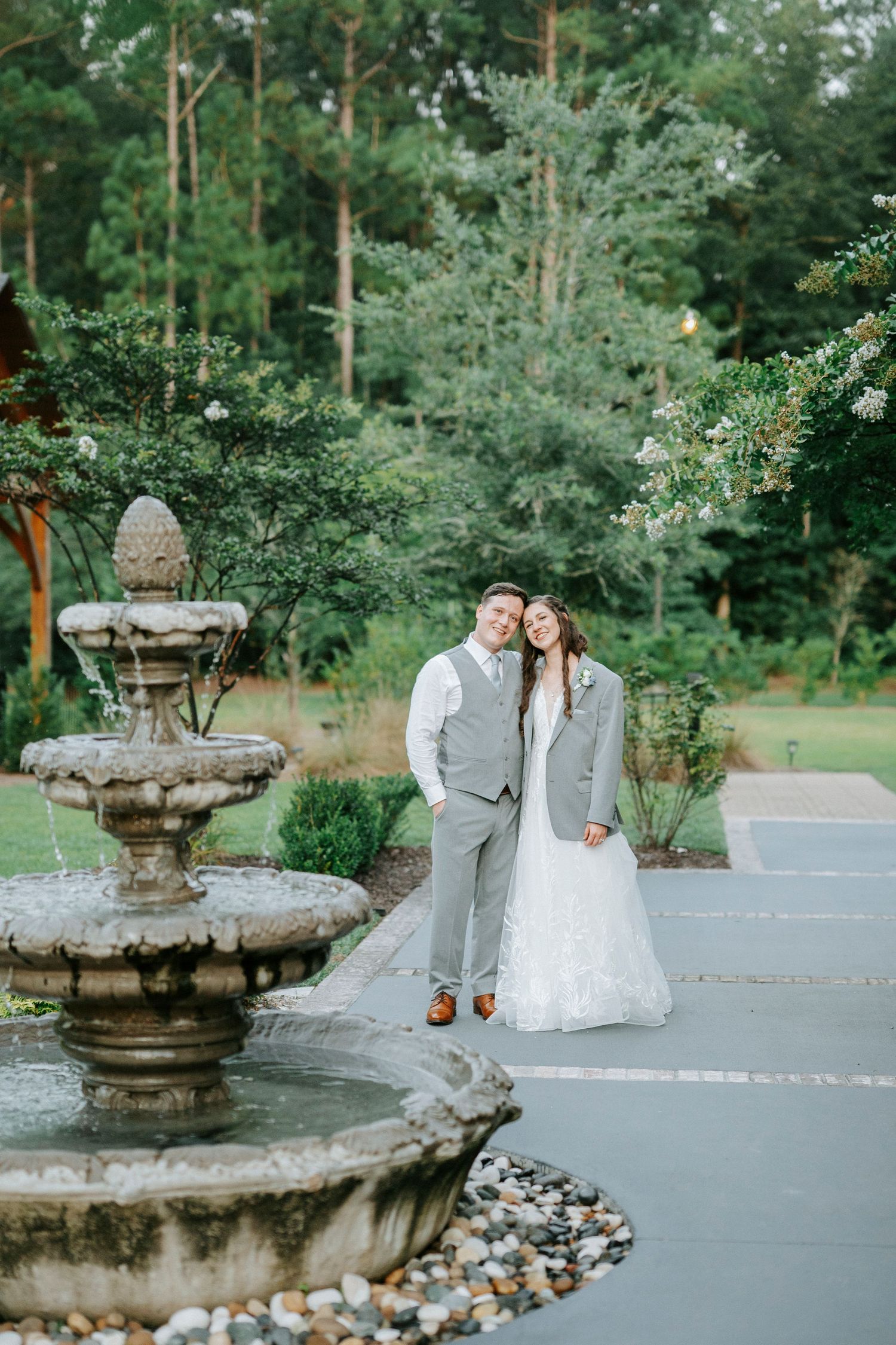 Romantic wedding couple poses near stone water fountains in a lush garden setting.