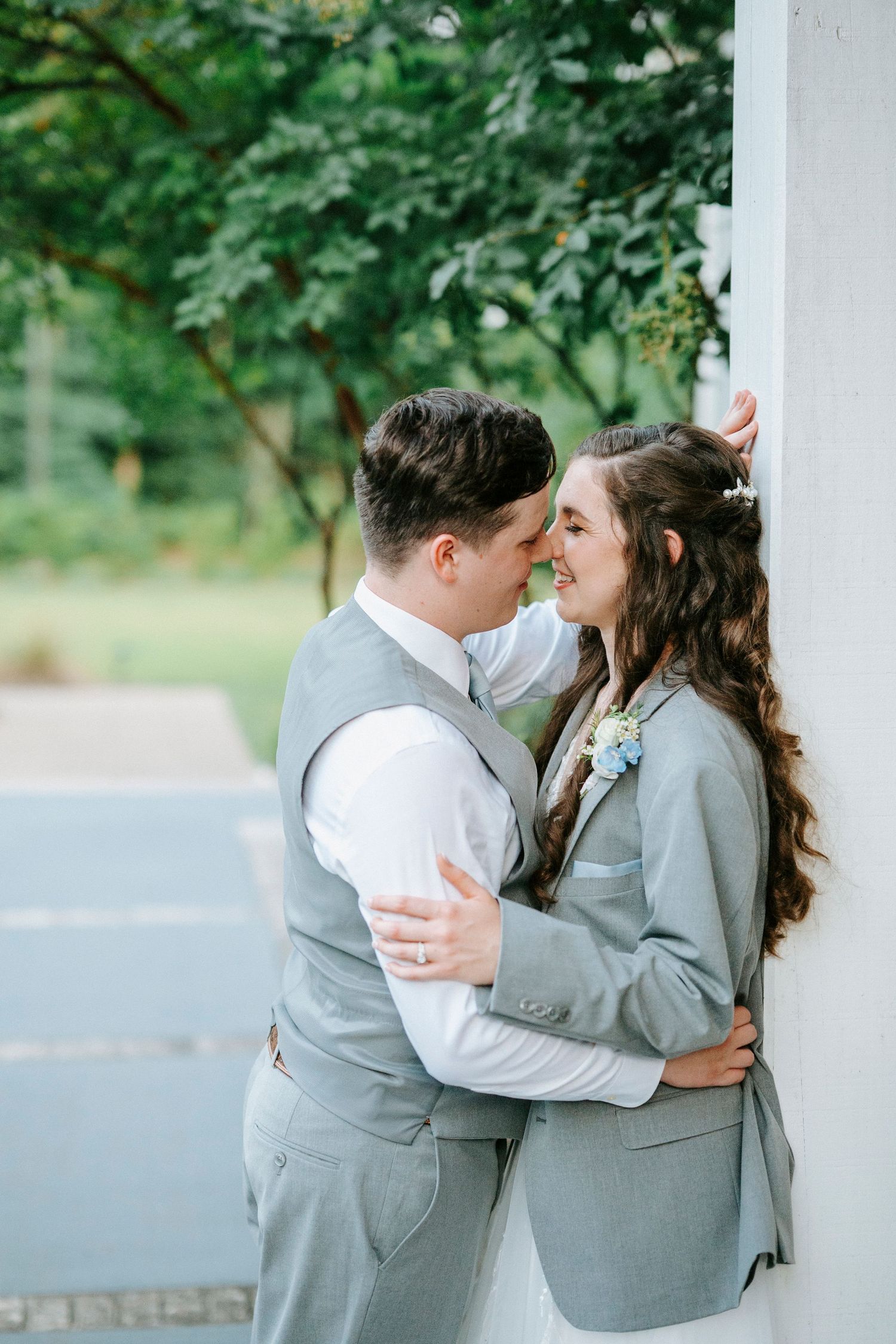 A romantic couple wearing matching gray suits embrace intimately outdoors with green foliage in the background.