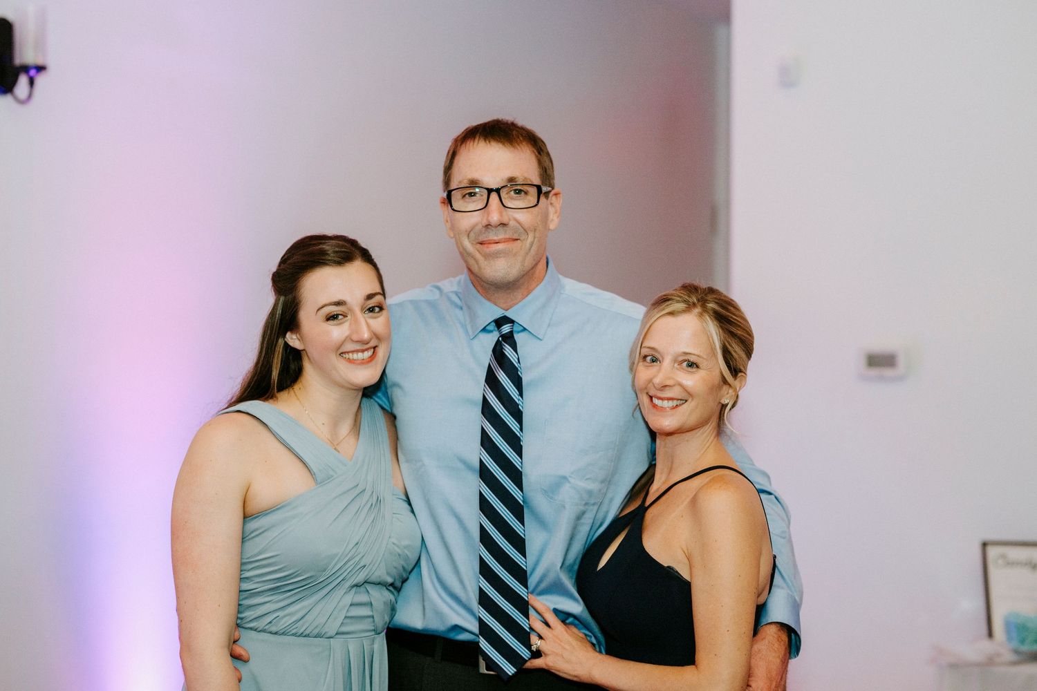 Wedding party members pose together in light blue attire against a white wall with purple lighting.