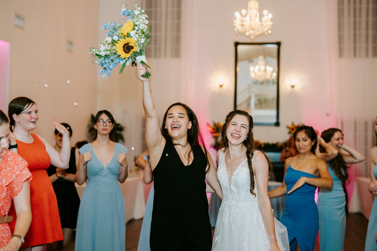 Wedding celebration with bridesmaids in blue dresses dancing and celebrating under elegant indoor lighting.