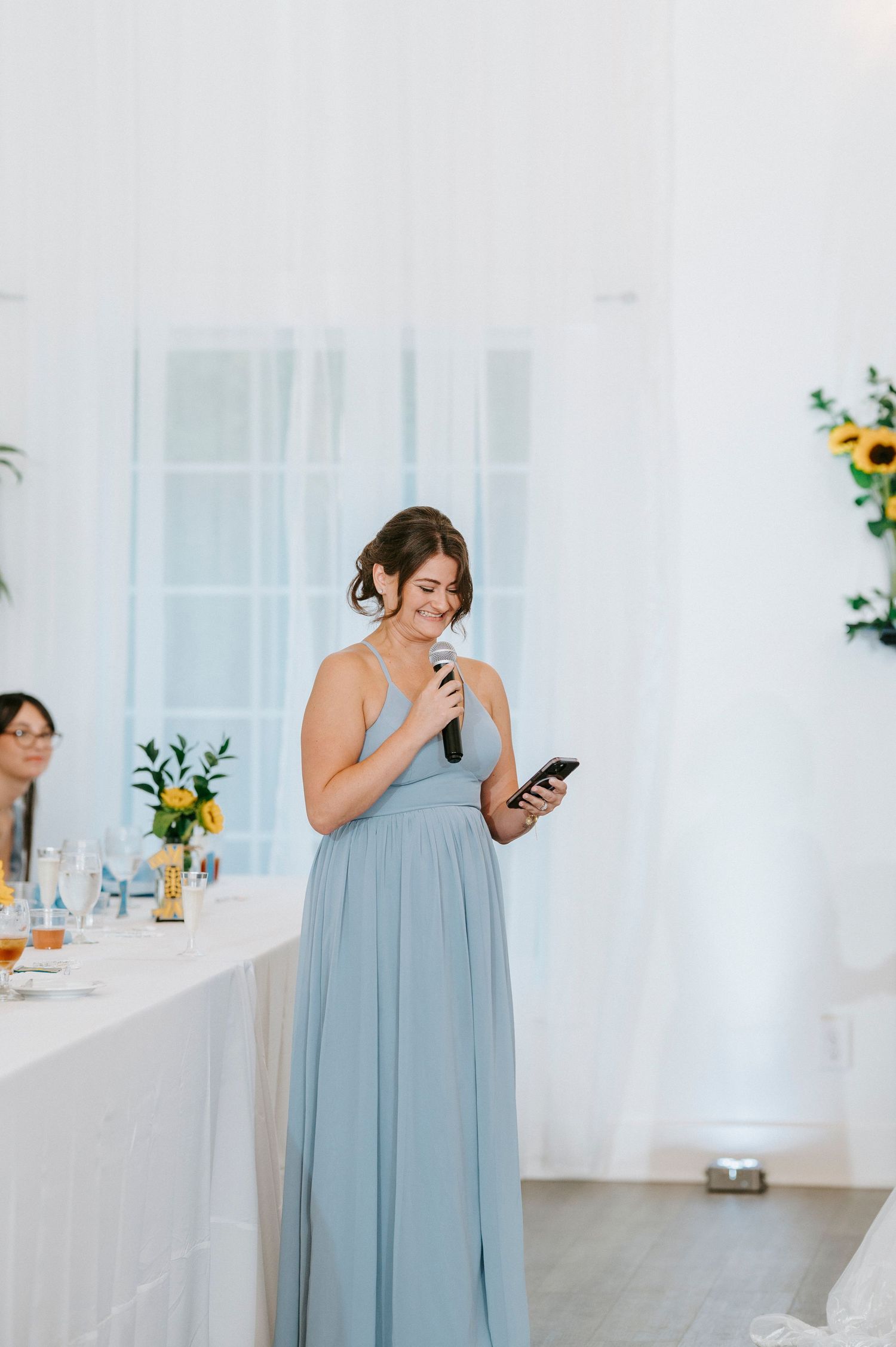 Wedding guest in dusty blue dress glances at phone while standing in bright white room with floral decorations.