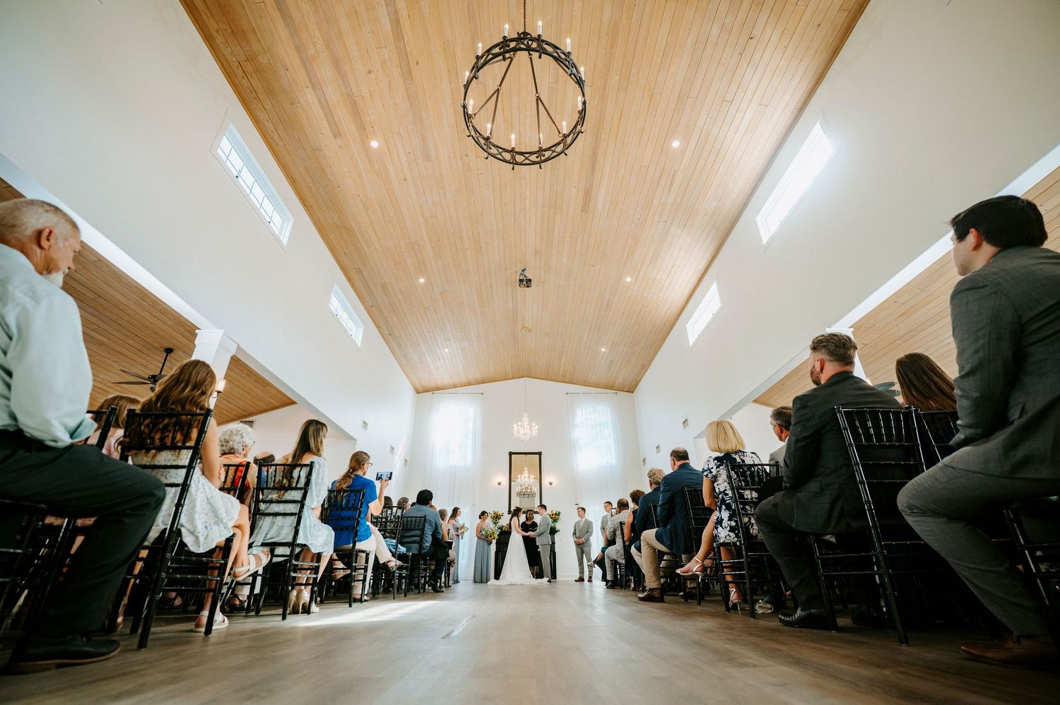 A wide-angle view of a wedding ceremony in a bright chapel with wooden ceiling beams.