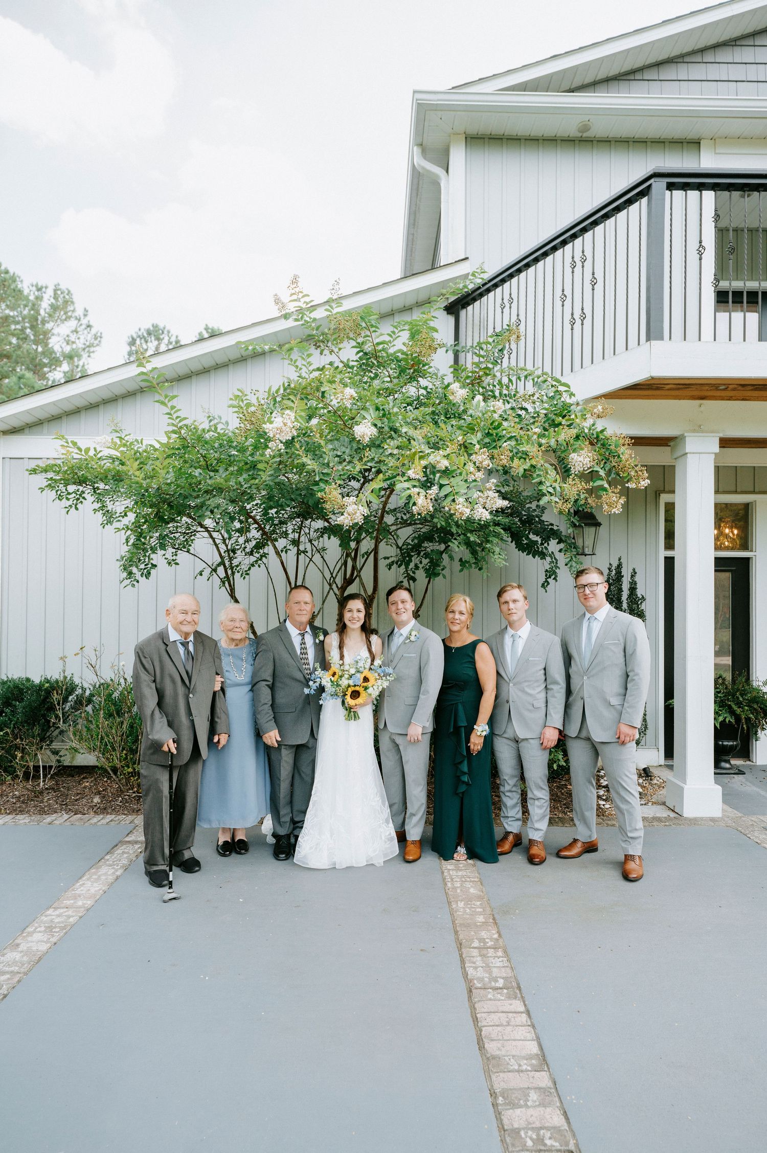 Wedding party in formal attire poses outside a white building with grey and white color scheme.