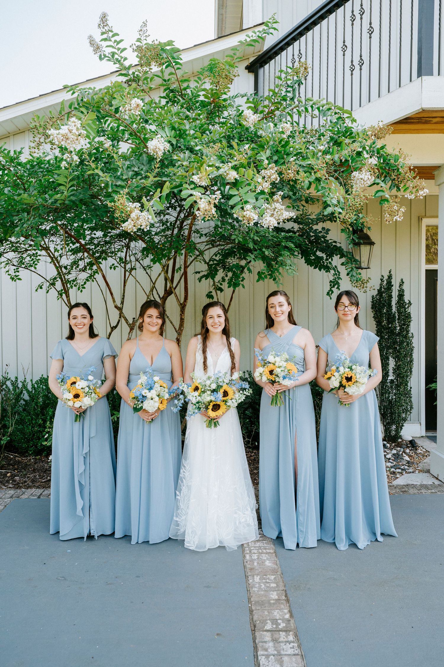 Wedding party in light blue bridesmaid dresses poses on a porch surrounded by greenery with vibrant sunflower bouquets.