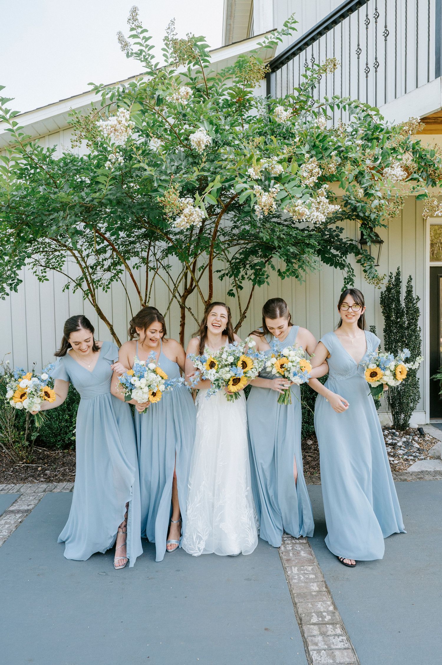 Bridal party wearing light blue gowns holding sunflower bouquets poses together outdoors.