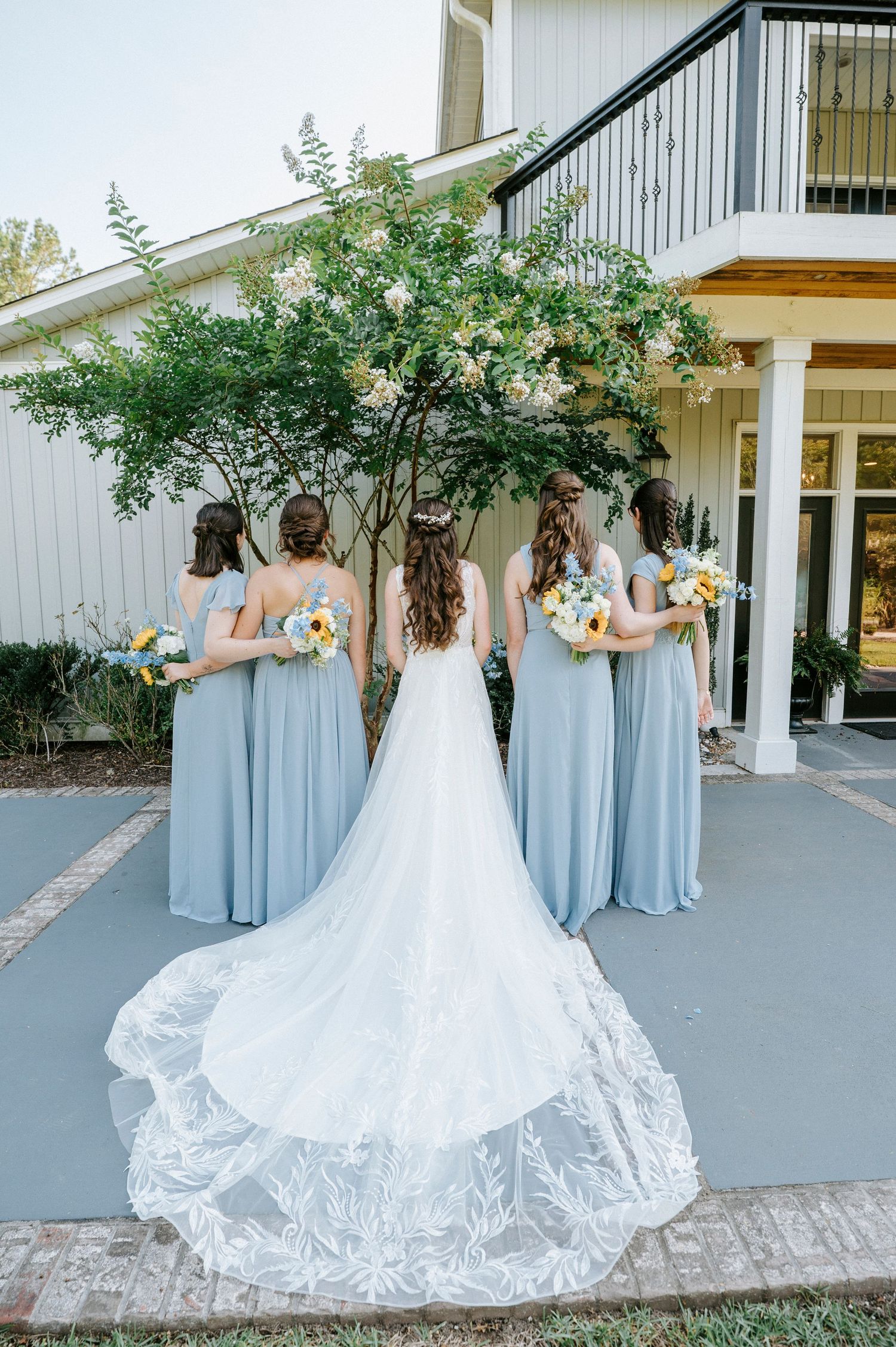 Back view of bridal party in dusty blue dresses with flowing white wedding gown train.
