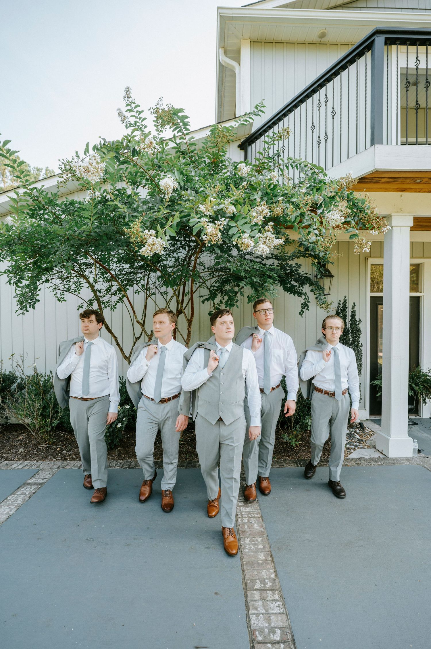Groomsmen in light grey suits and brown shoes pose together outside a white building.