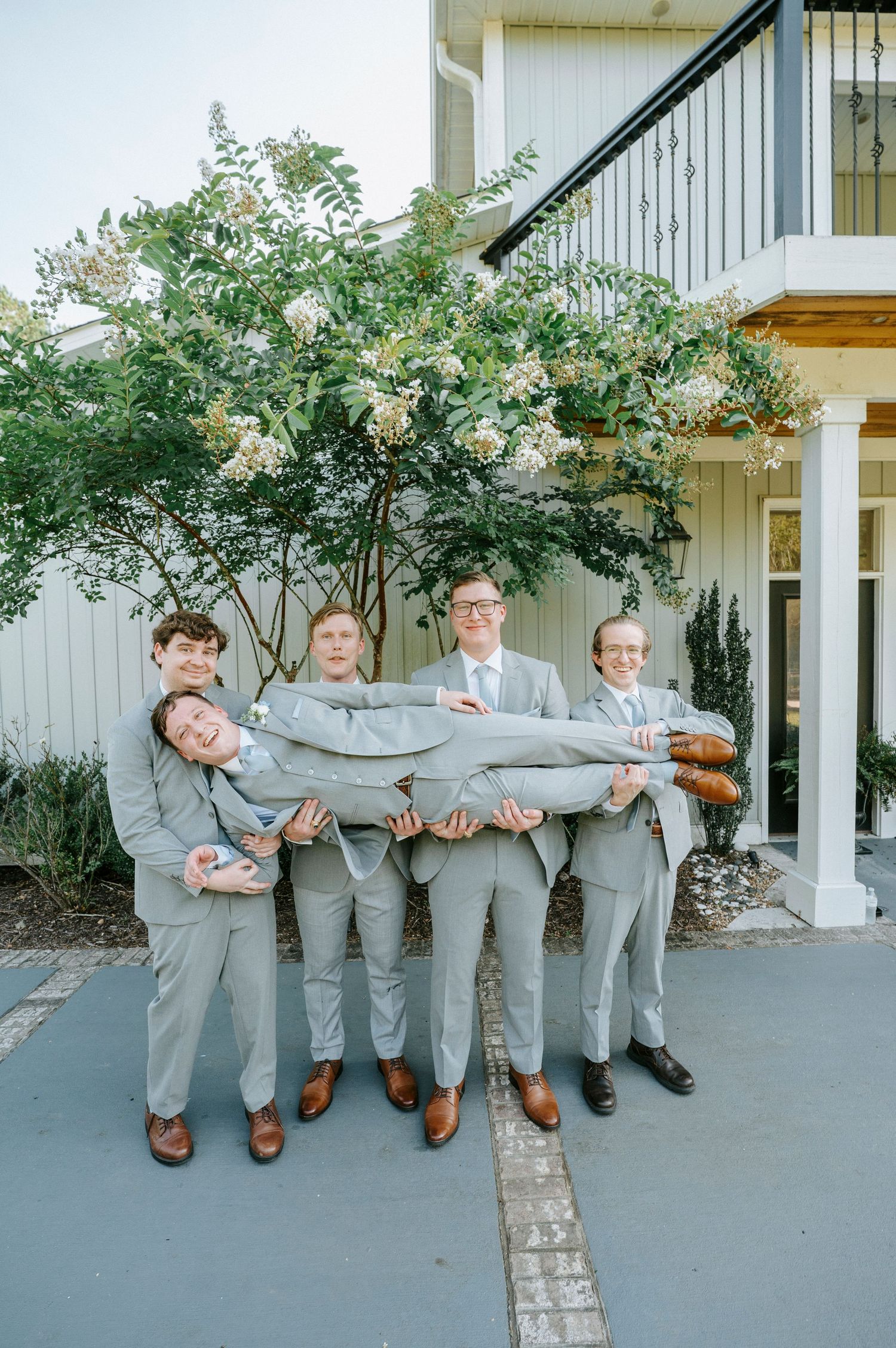 Groomsmen in matching grey suits strike playful poses against white building exterior.