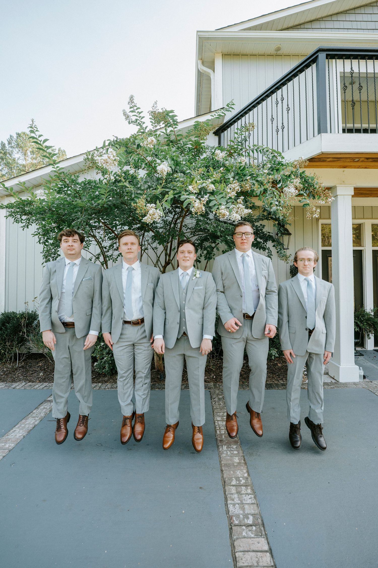 A group of groomsmen in light gray suits and brown shoes stand lined up outdoors against a white building.