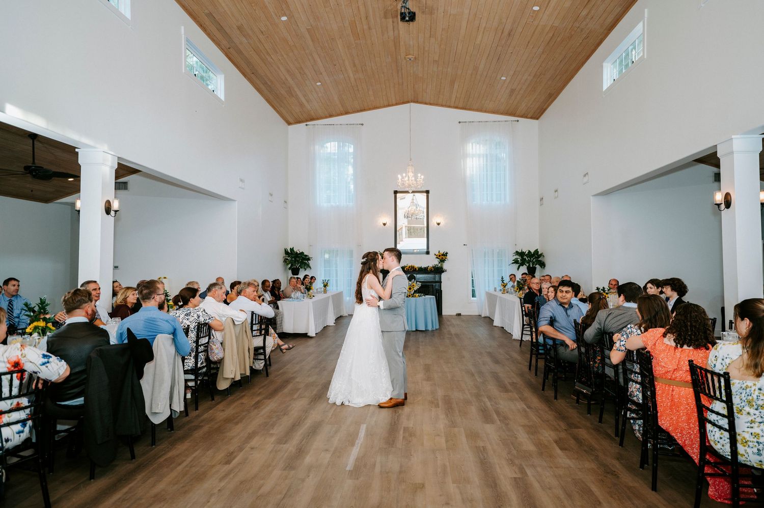 Wedding guests dance and celebrate in a modern white reception venue with wooden ceiling beams and uplighting.