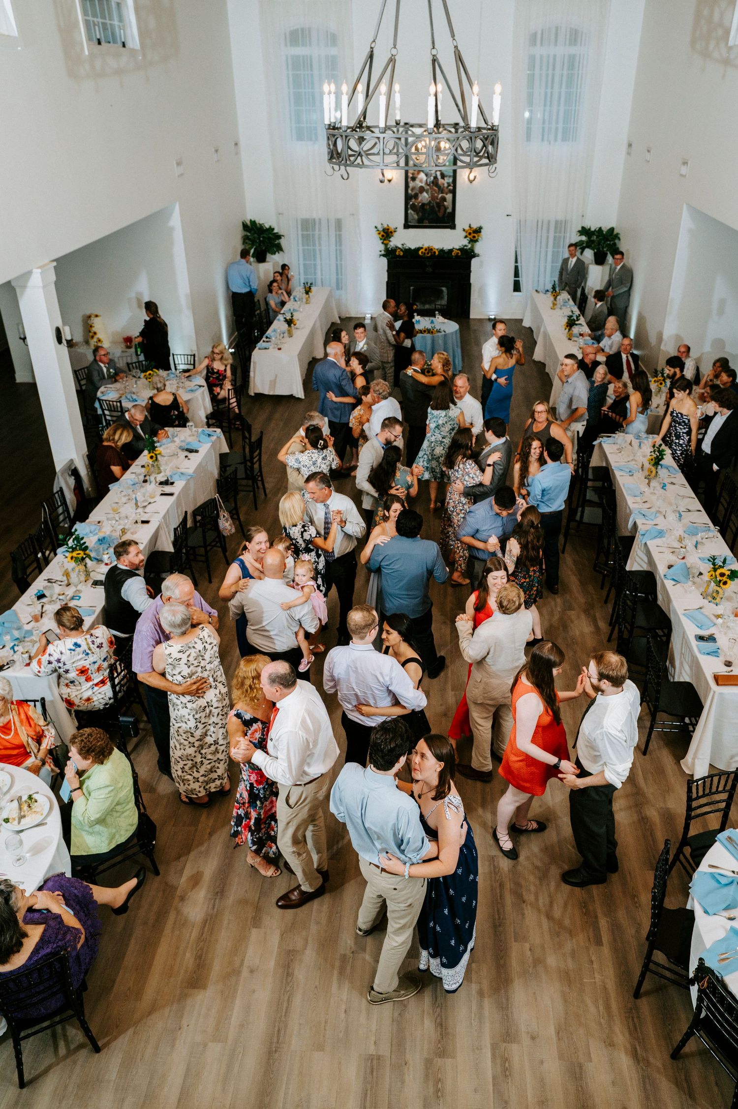 Wedding reception venue with guests mingling and dining at long tables in an elegant white room with chandeliers.
