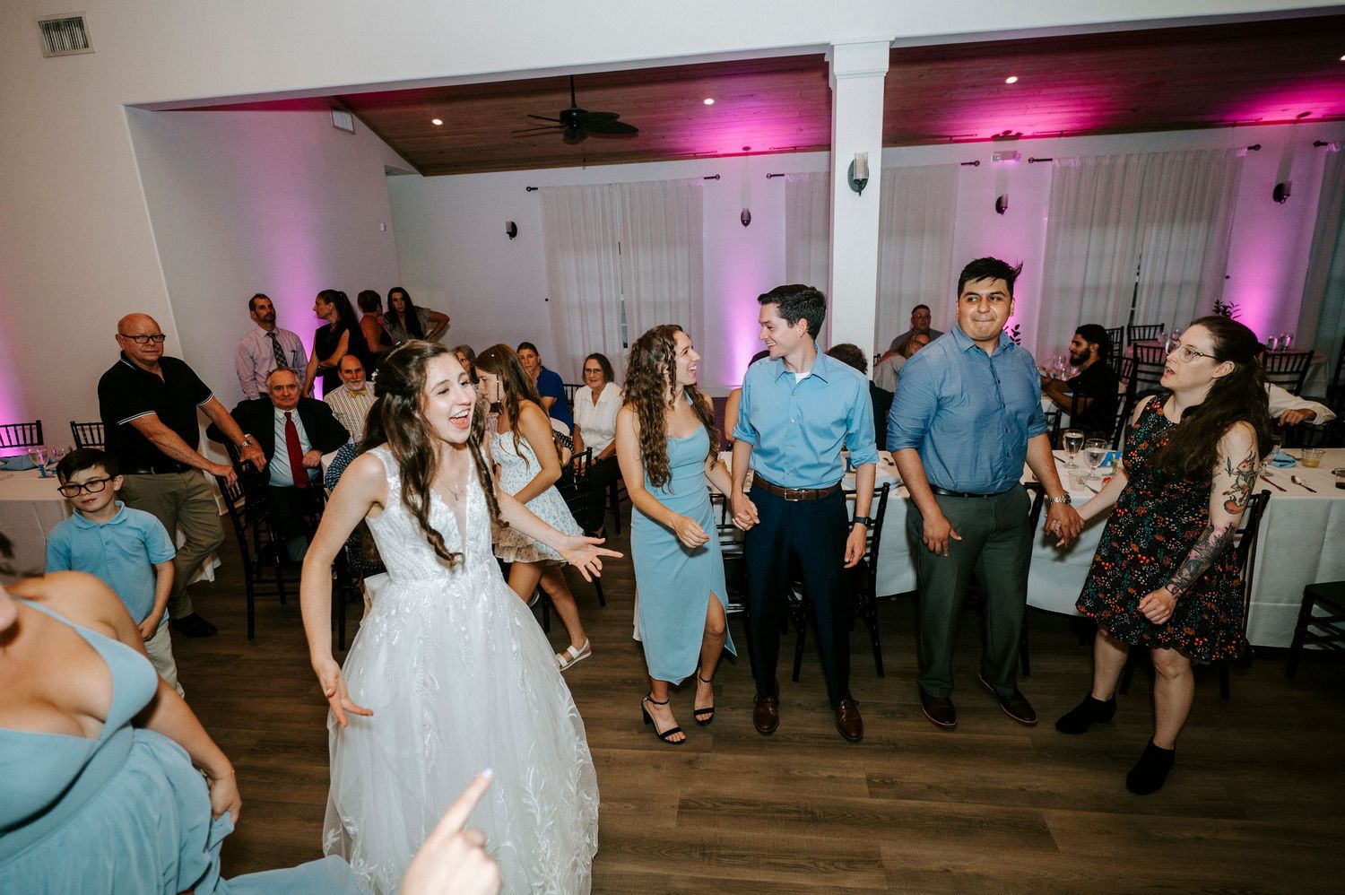 Wedding guests fill a modern venue dance floor illuminated by pink and purple lighting.