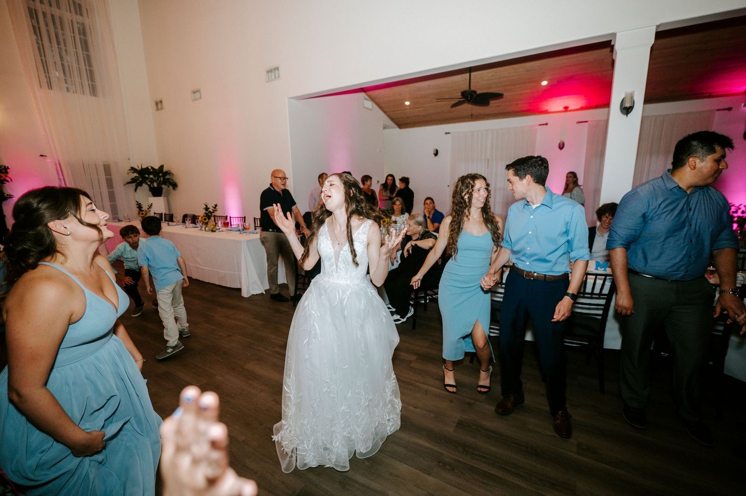 Wedding reception dance floor filled with guests dancing under pink mood lighting and white walls.