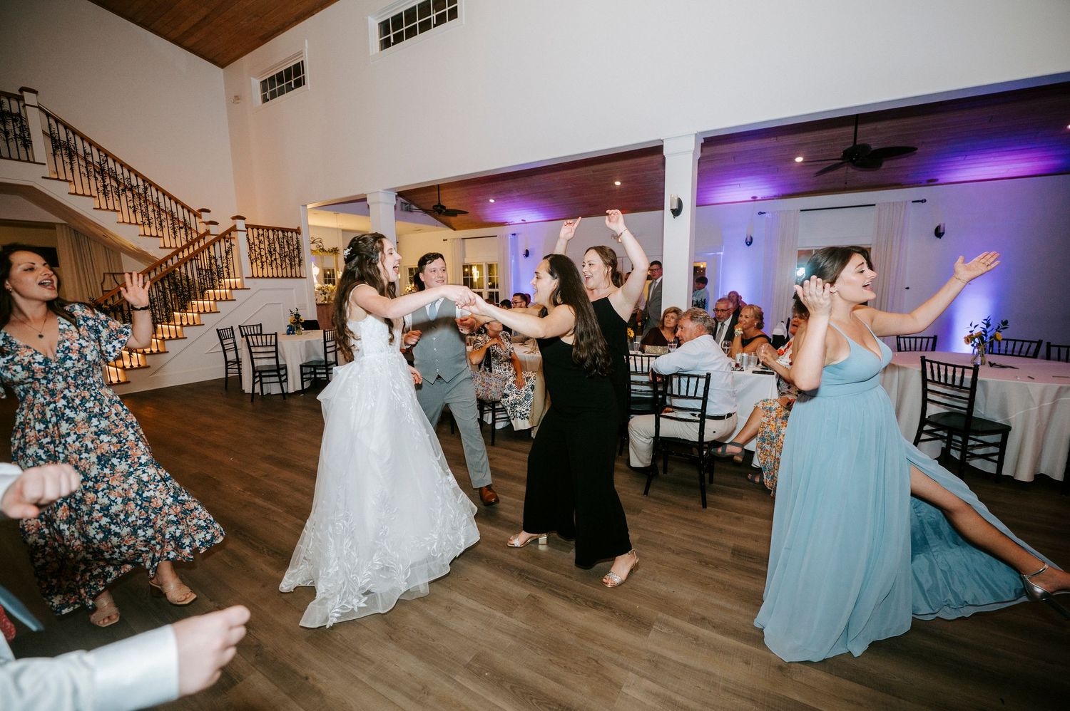 Wedding guests dancing and celebrating on a wooden dance floor with purple uplighting at a reception venue.