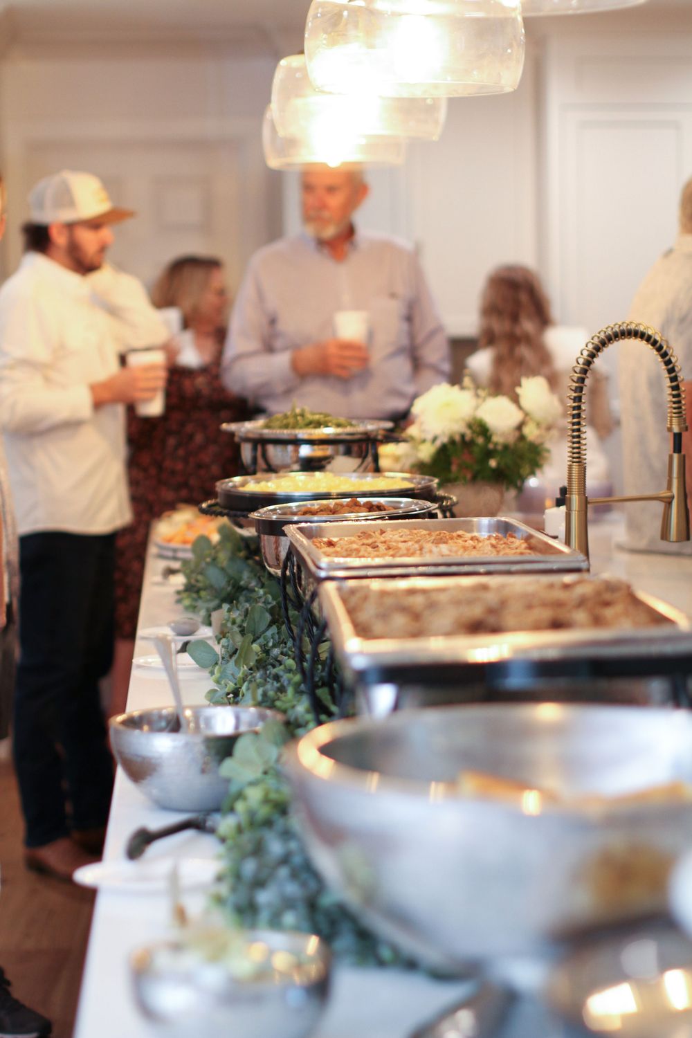 Catering staff serve food at an elegant buffet line with stainless steel chafing dishes and decorative greenery.