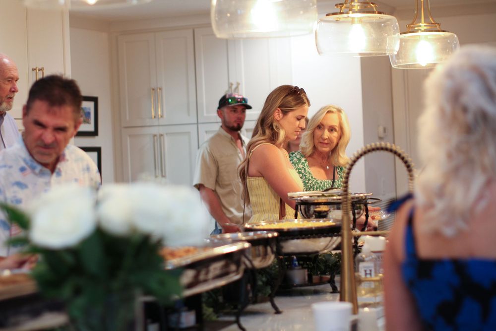 A group gathers around a buffet station at an indoor event with pendant lighting overhead.