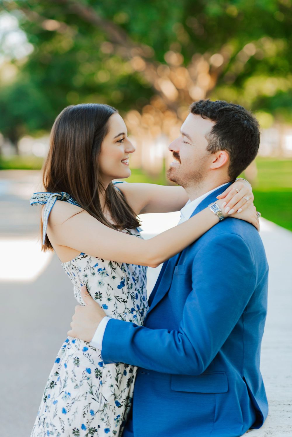 A couple in a floral dress and blue suit share a loving embrace in an outdoor garden setting.