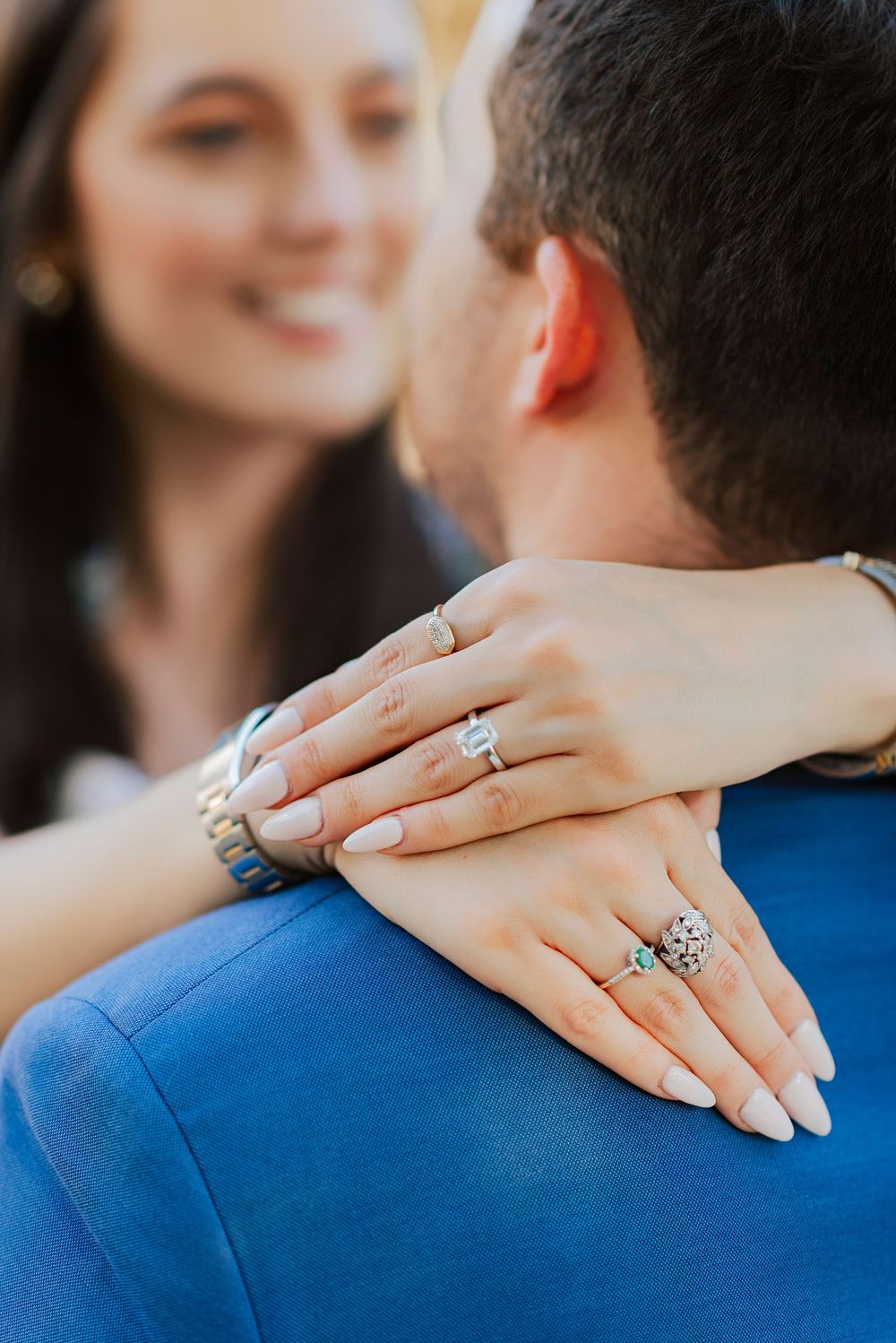 Close-up detail shot of hands wearing multiple rings and jewelry against a blue blazer.