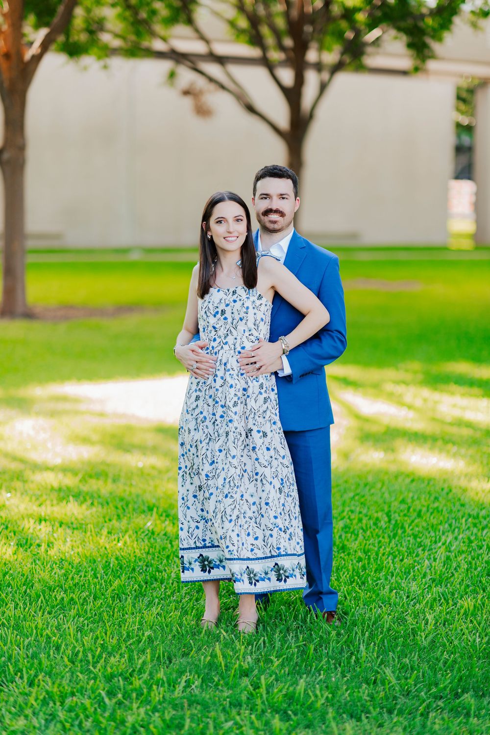 A couple poses outdoors in coordinated outfits with a blue suit and floral maxi dress against a grassy background.