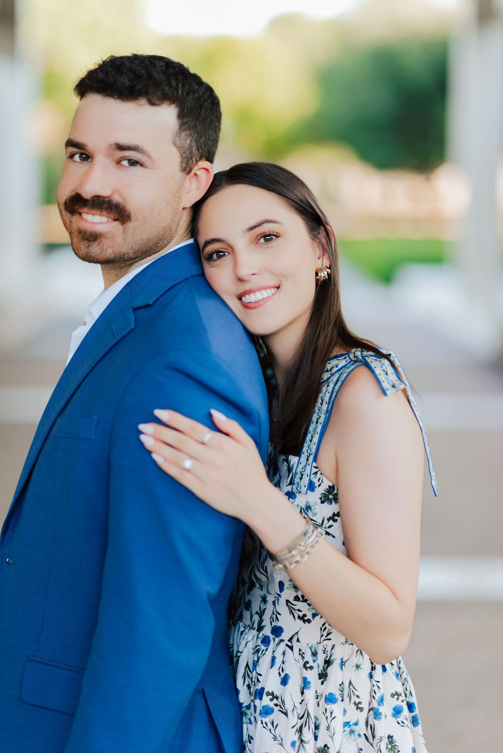 A couple in coordinating outfits pose together, with bright smiles and a blue suit against light architecture.