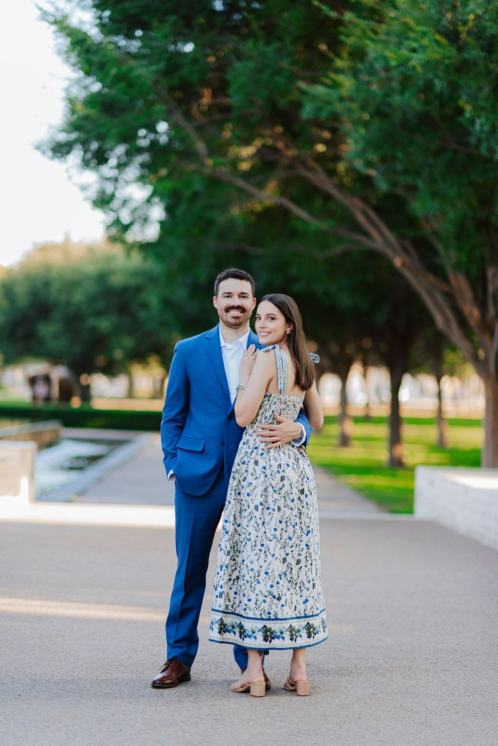 A couple in formal wear embraces on a pathway with trees in the background during golden hour.