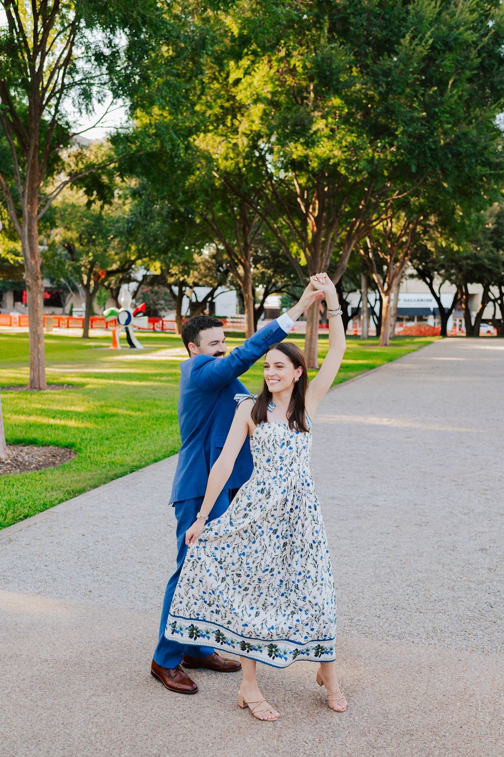 Couple dancing playfully in a park with green trees, wearing a blue suit and floral maxi dress.