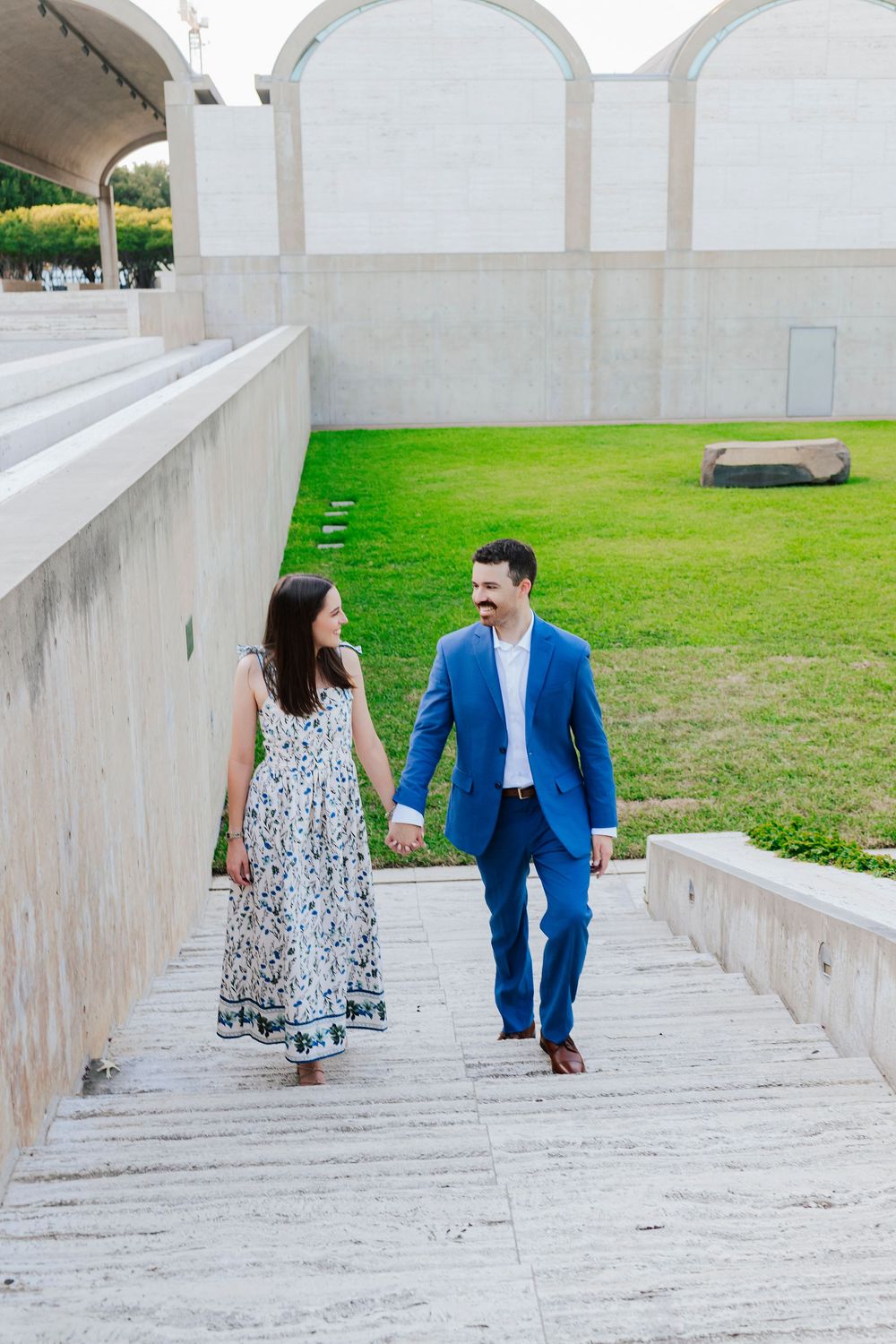 A couple walks hand in hand up concrete steps with arched architecture and green lawn.