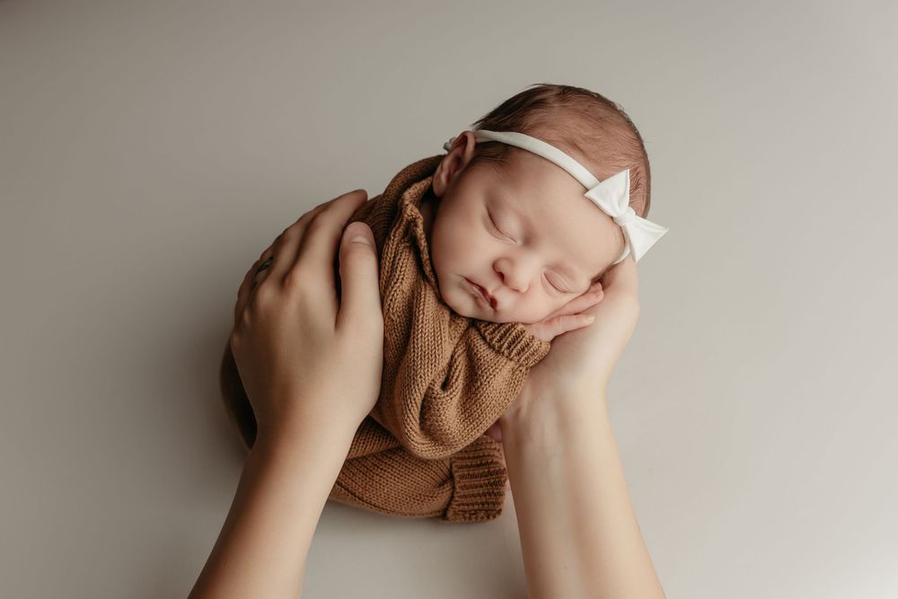 newborn girl in brown jumper, posed in mama's hands, taken by Jen Knightstep, Photographer