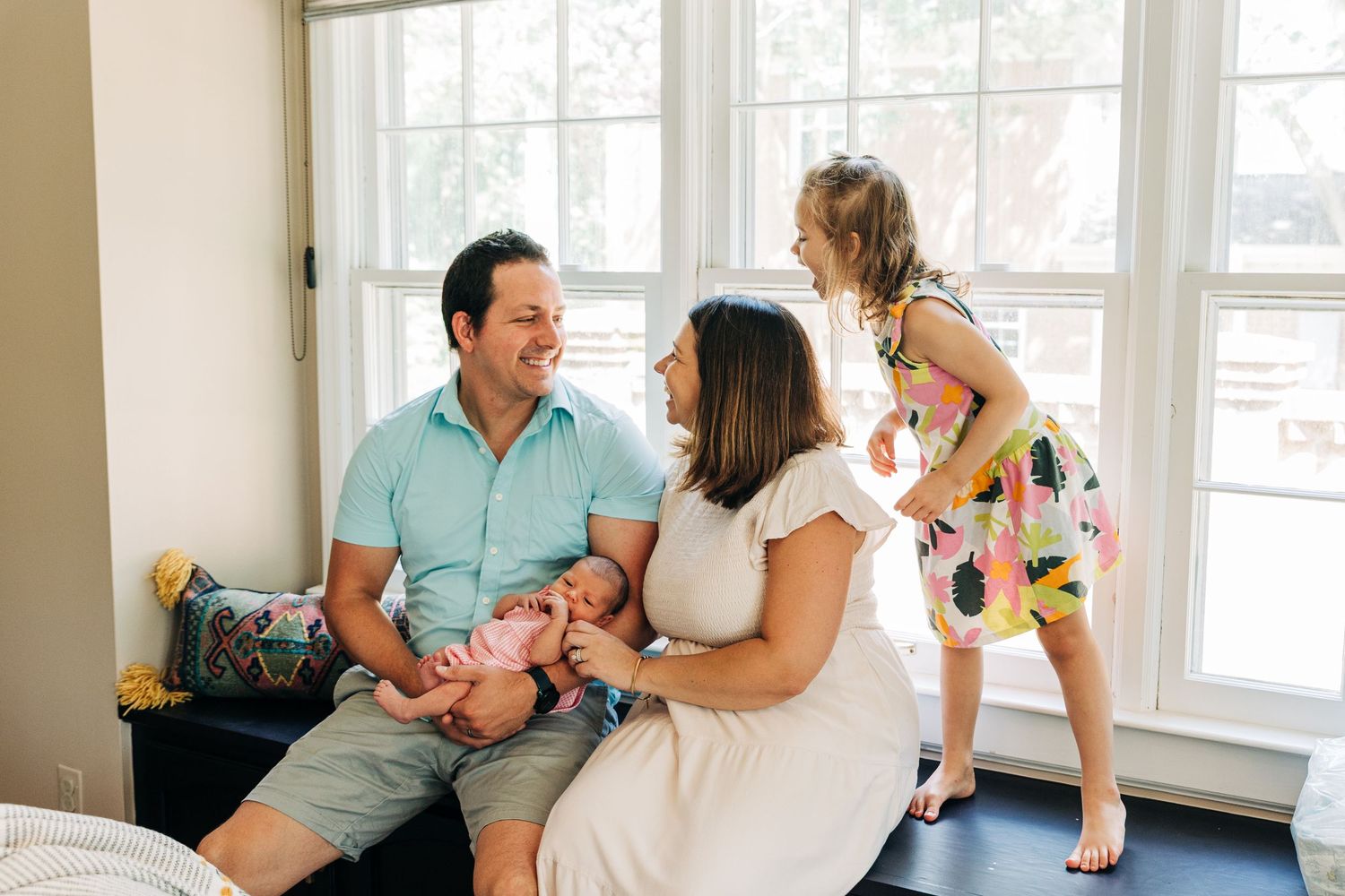 A happy family lounges together in a bright sunlit room by large windows on a cozy couch.