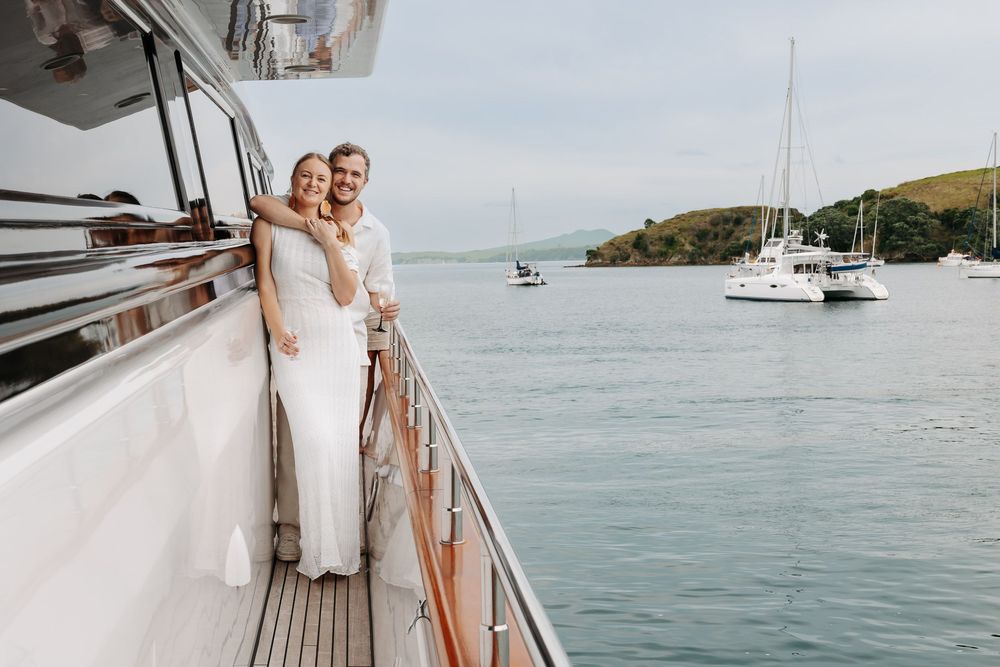 A couple in elegant white attire stands on a yacht deck overlooking a scenic harbor with sailboats in the background.