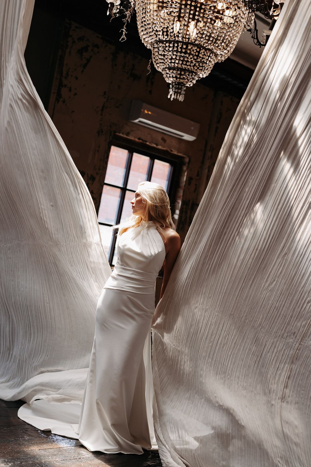 A bride in a flowing white gown stands elegantly beneath crystal chandeliers as her veil billows in a dramatic sequence.