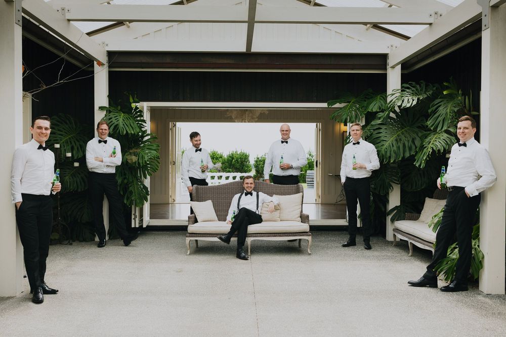 Wedding party stands in a covered outdoor walkway with white columns and green foliage in the background.