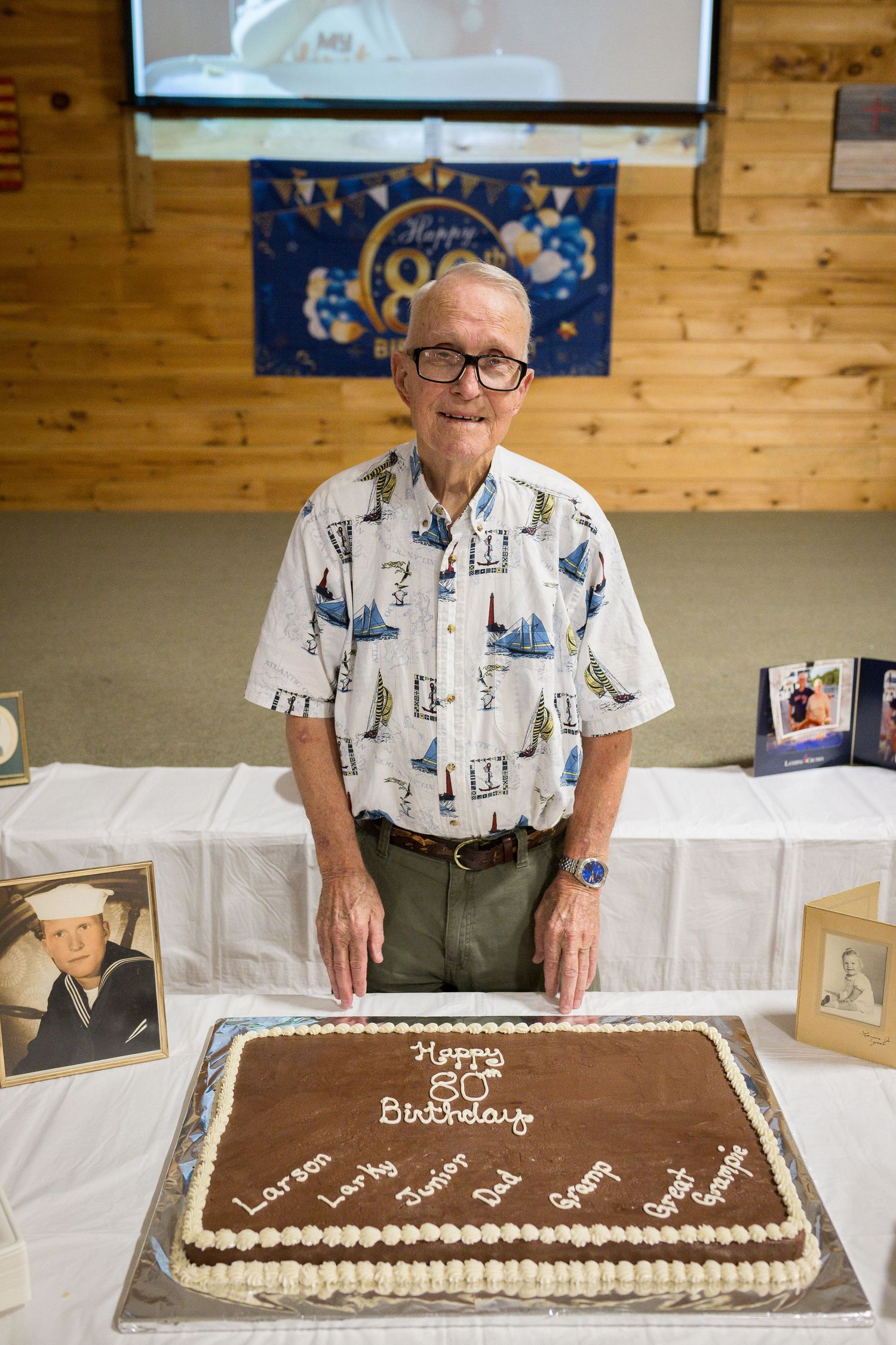 Large chocolate birthday cake with white frosting border displayed on a table with wood paneled walls.