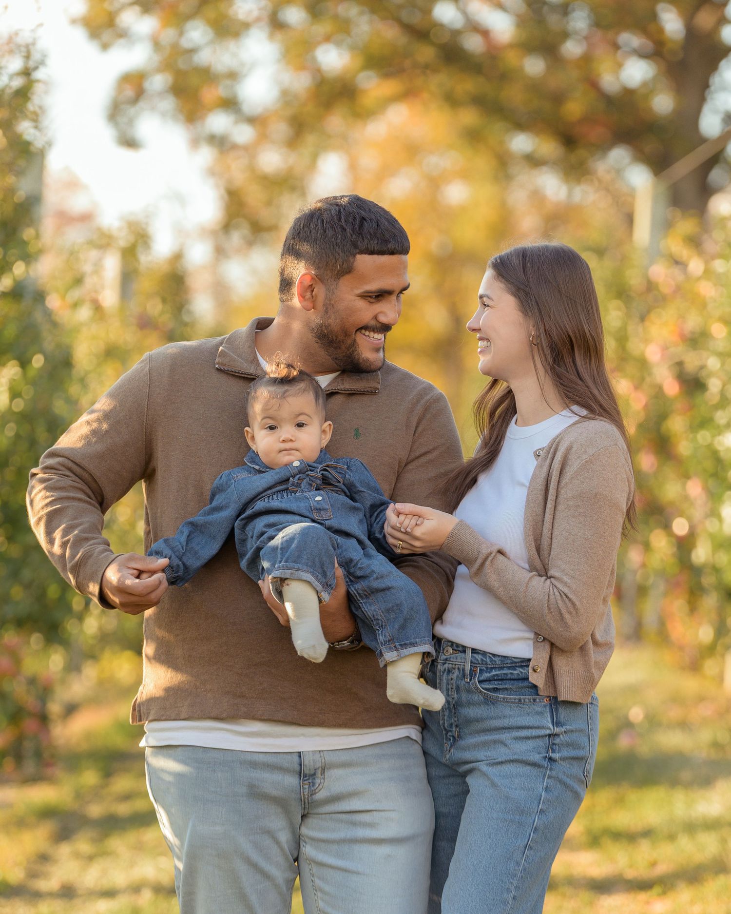 Family wearing brown and white outfits poses together during autumn photoshoot in park setting.