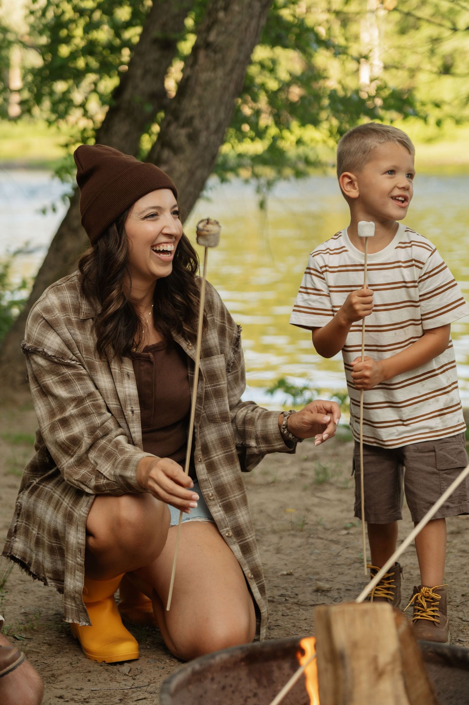A woman and young boy roast marshmallows over a campfire by a lake, enjoying a fun outdoor camping experience.
