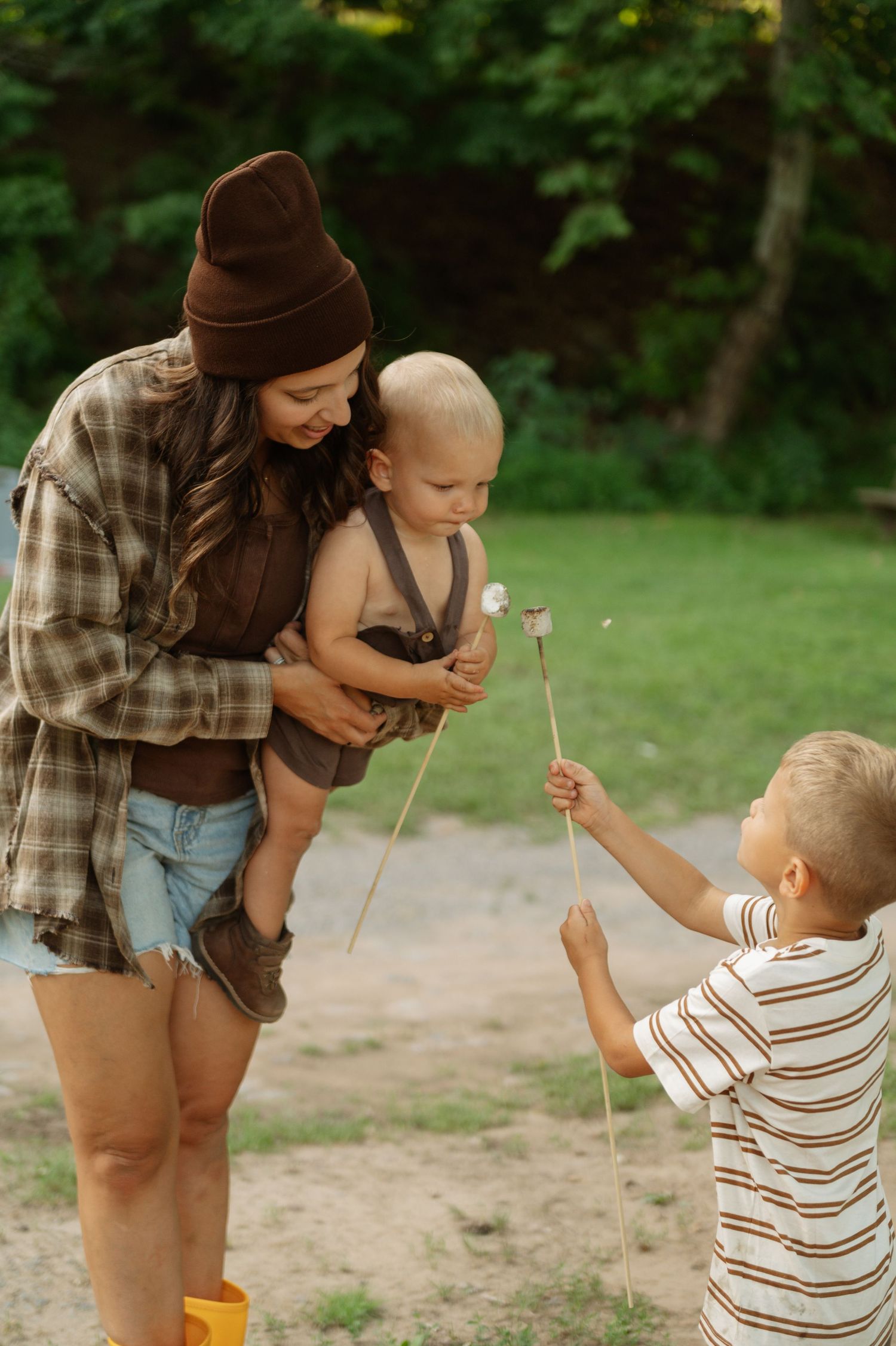 A mother holding a baby while an older boy shows them dandelions outdoors in a lush green park setting.