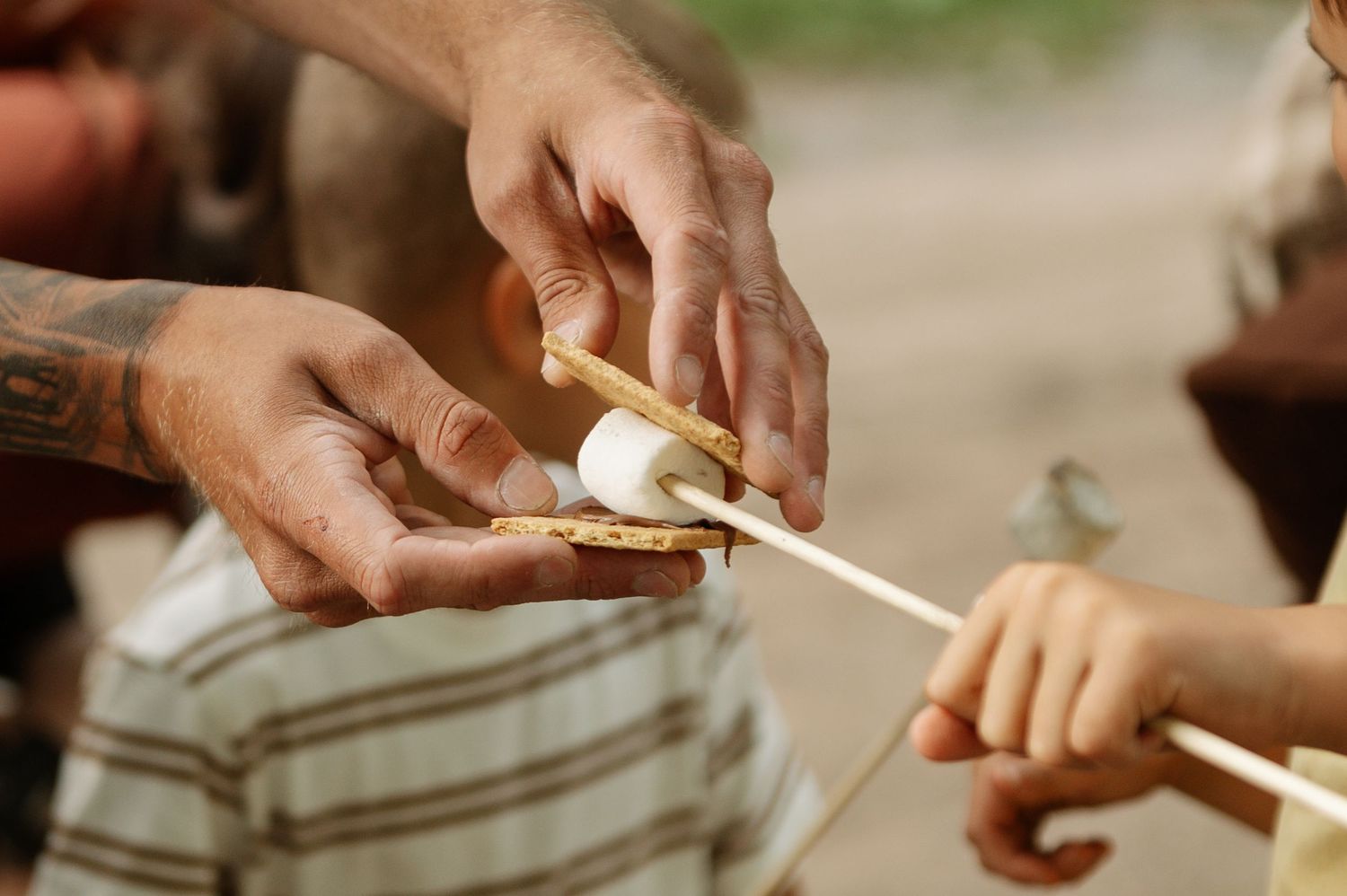 Tattooed adult hands helping a child roast a marshmallow on a stick over a campfire outdoors.