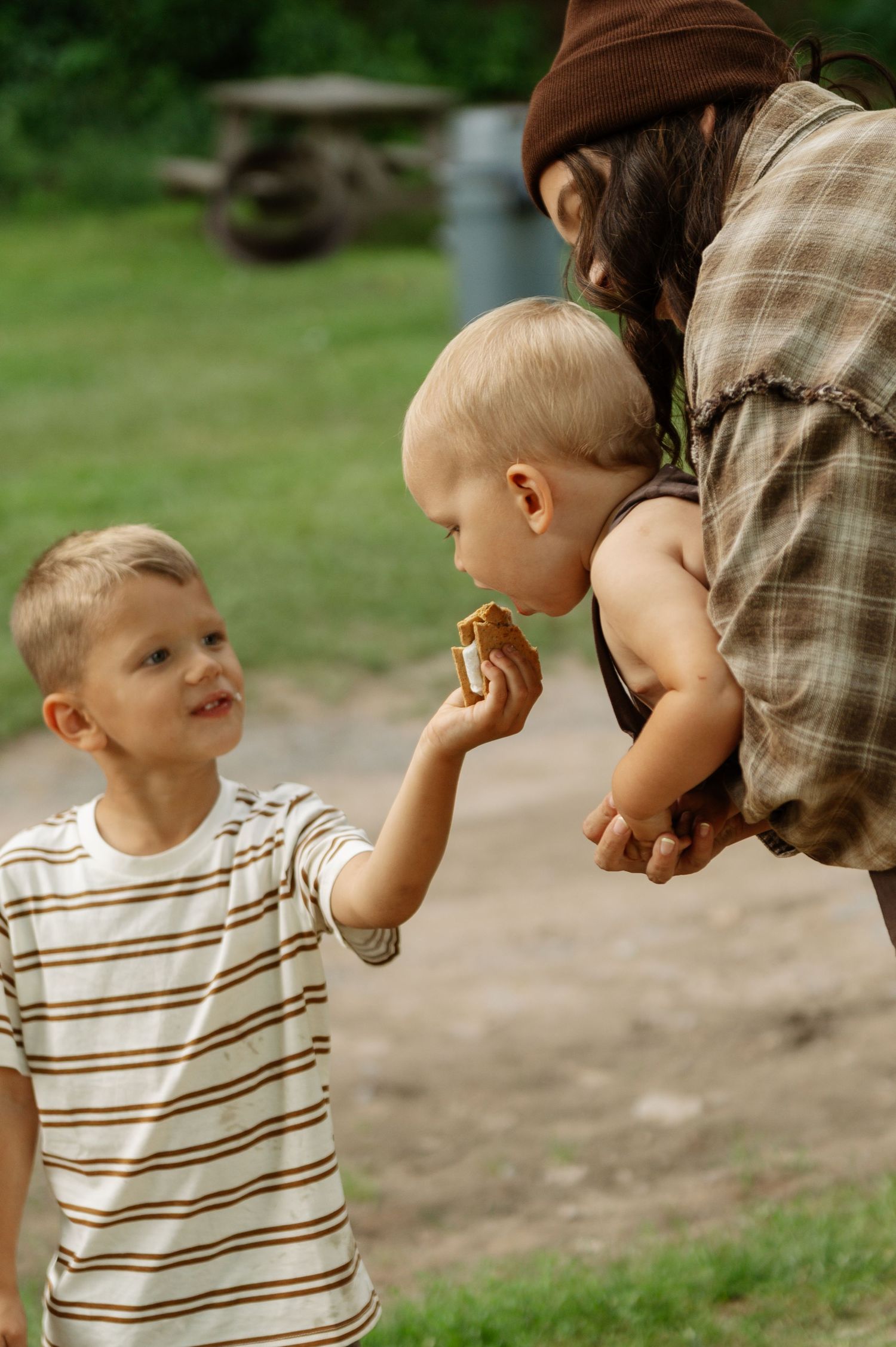 Two young boys outdoors sharing a snack, with an adult woman in a plaid shirt watching them in a green park setting.