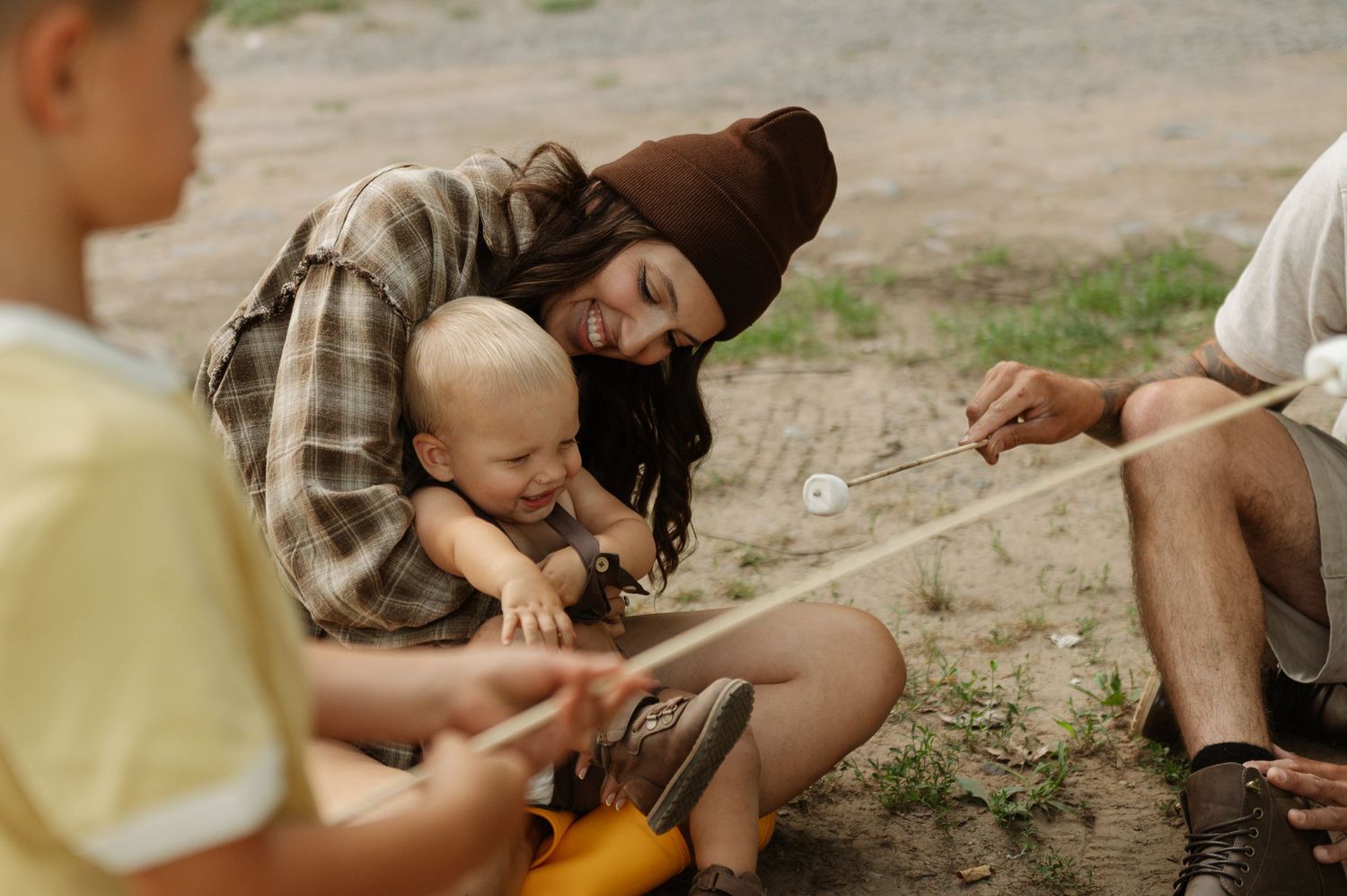A parent in a beanie hat holds a baby while roasting marshmallows with children around a campfire outdoors.