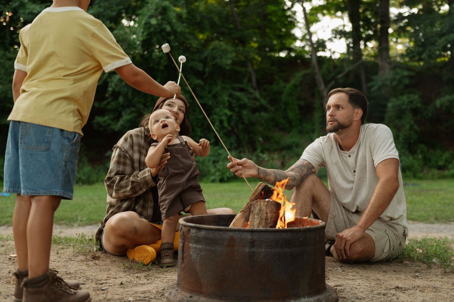 Family roasting marshmallows over a campfire outdoors, surrounded by trees on a warm summer day.