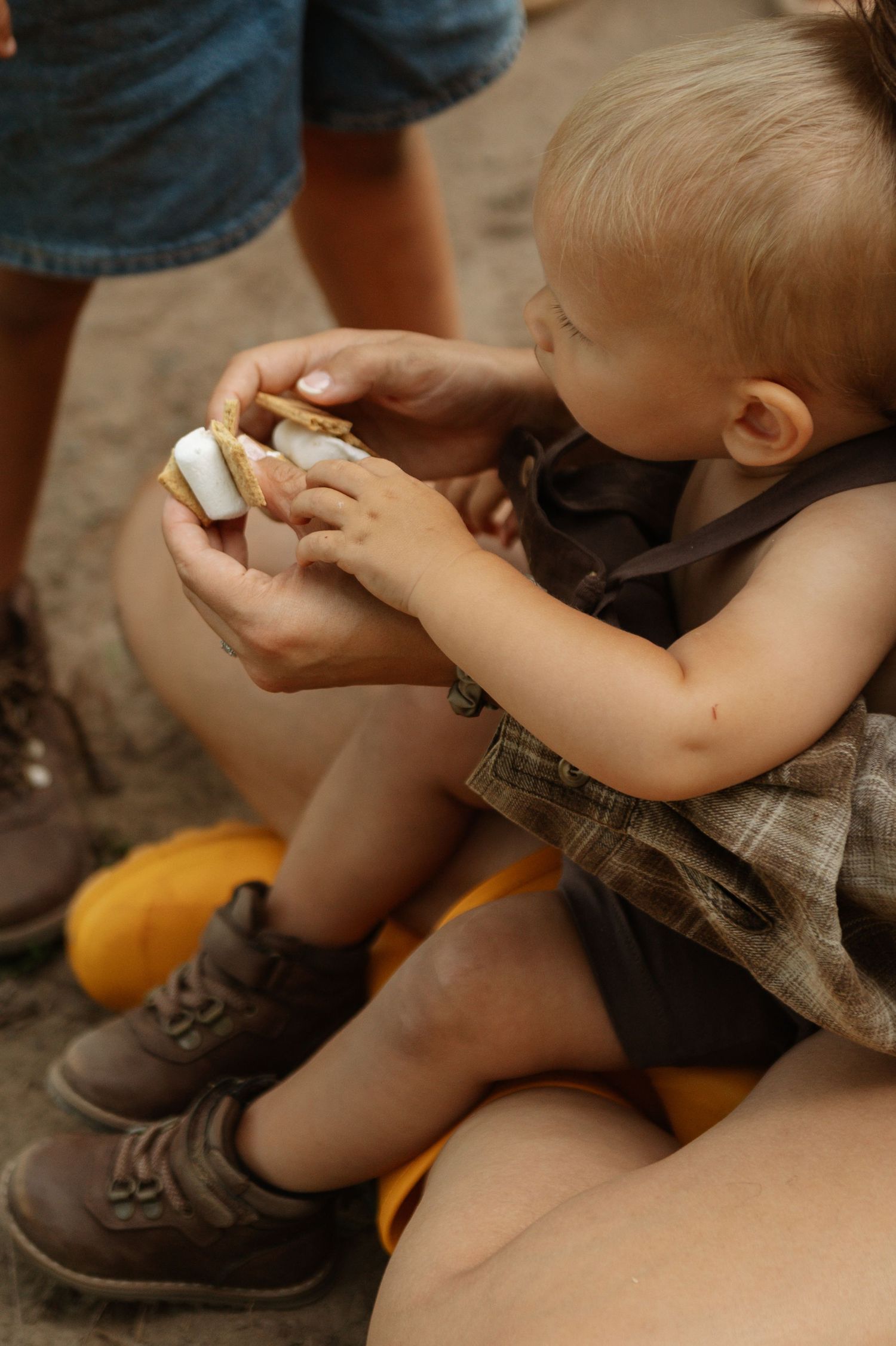 A toddler in a plaid romper eagerly eats a s'more held by an adult outdoors.